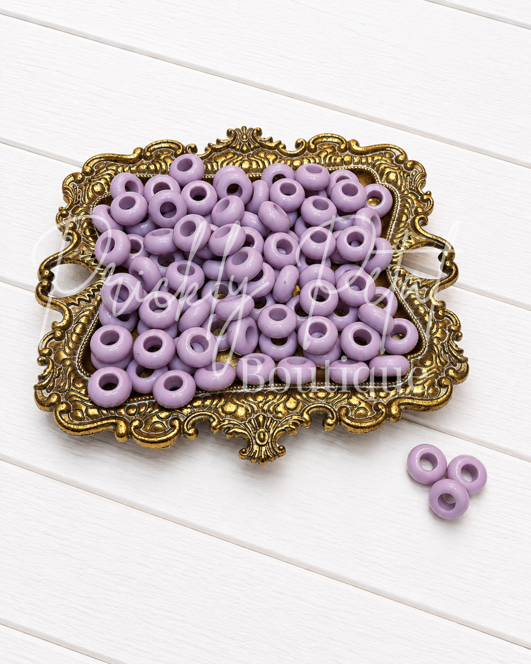 Lavender purple flatback seed beads arranged on an ornate gold-colored tray, with three additional beads placed on the white wooden surface beside the tray.