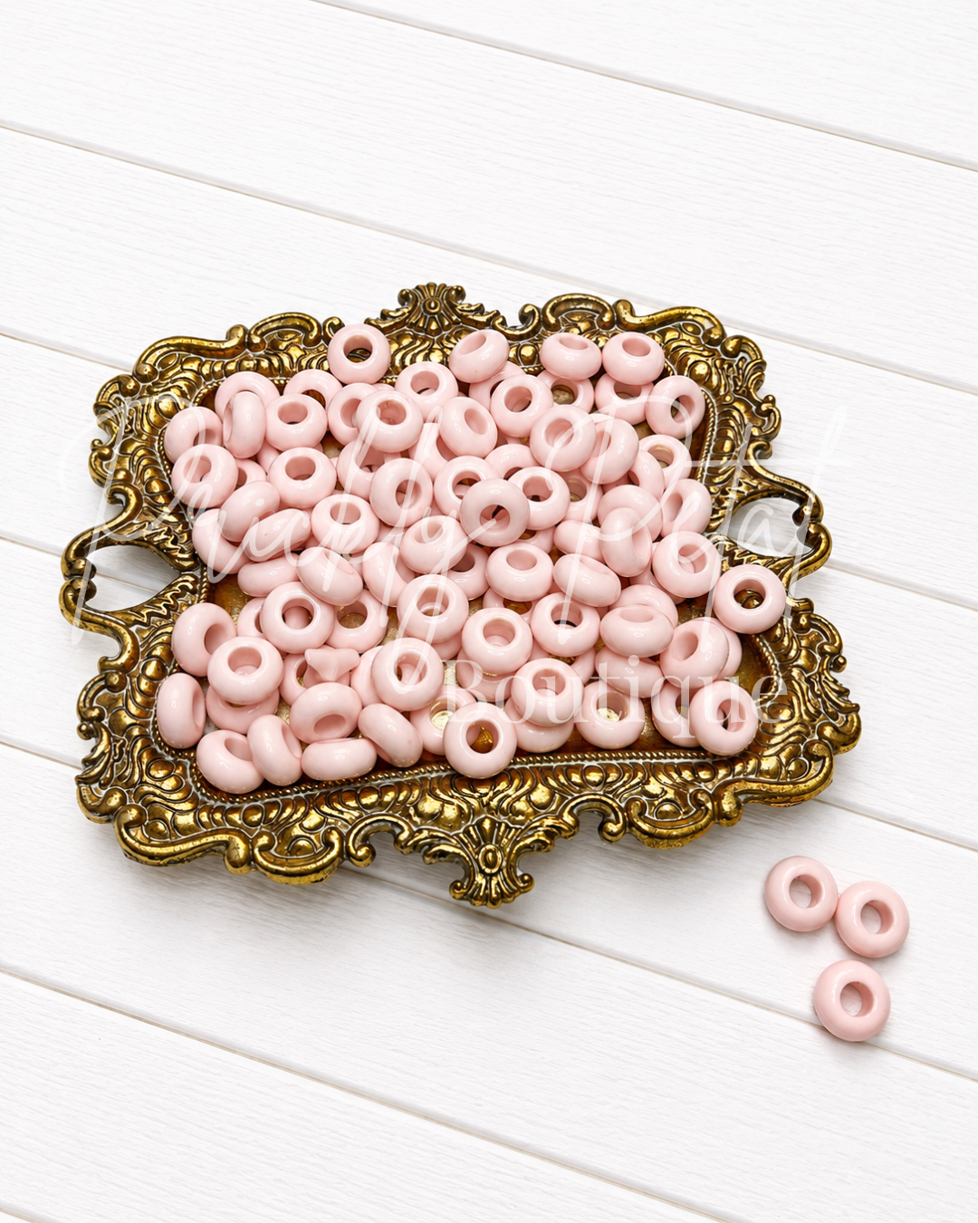 Light pink round spacer beads with a central hole, displayed on an ornate gold-colored tray over a white wooden background.