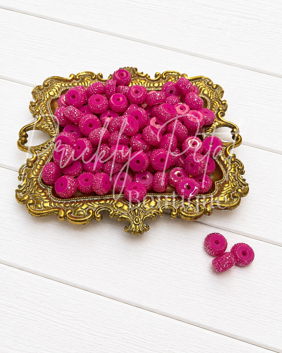 Bright pink glittery flatback craft beads piled in an ornate gold tray on a white wood background, with three loose beads in the lower right