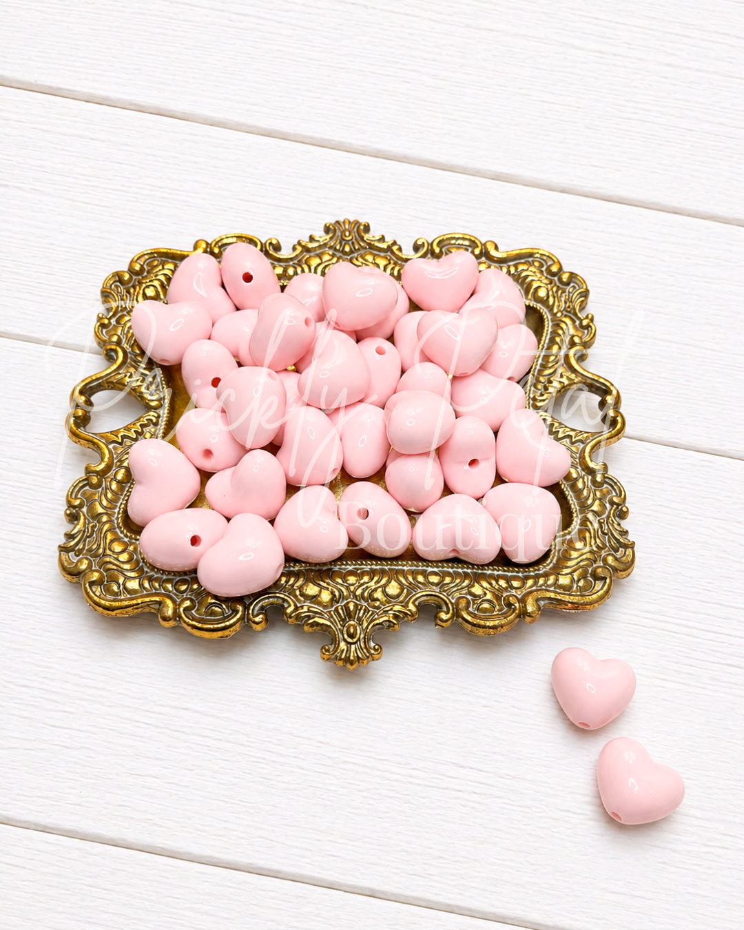 Pink heart-shaped craft beads with holes, displayed on an ornate gold-colored tray with two additional hearts on a white wooden surface.