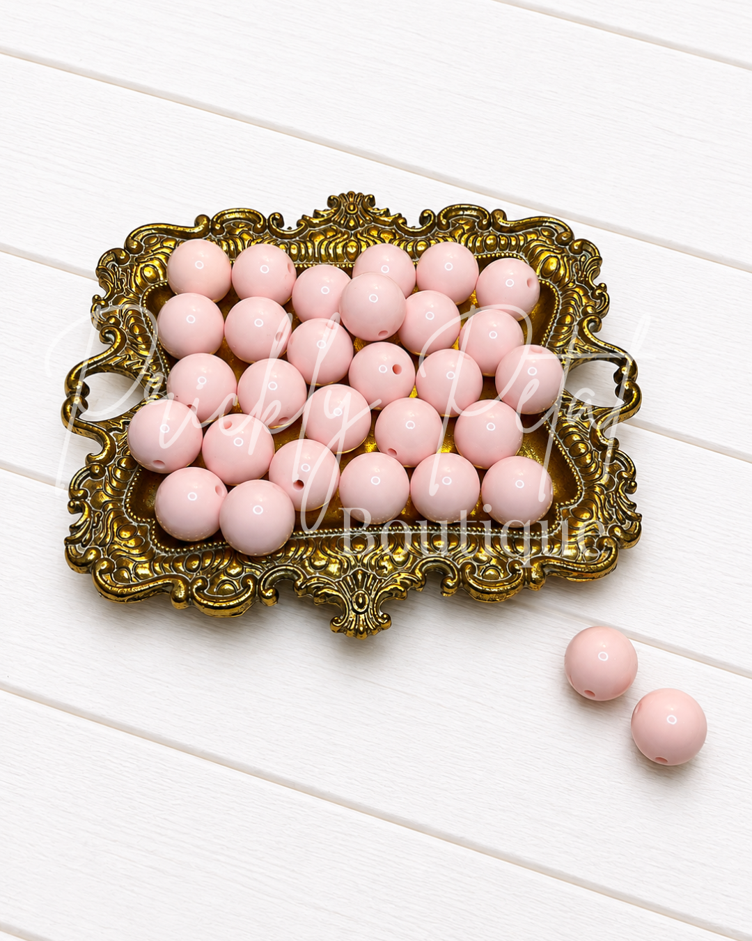 Light pink round glass beads with pre-drilled holes, displayed in an ornate gold-colored metal tray on a white wooden background, with two beads placed outside the tray.
