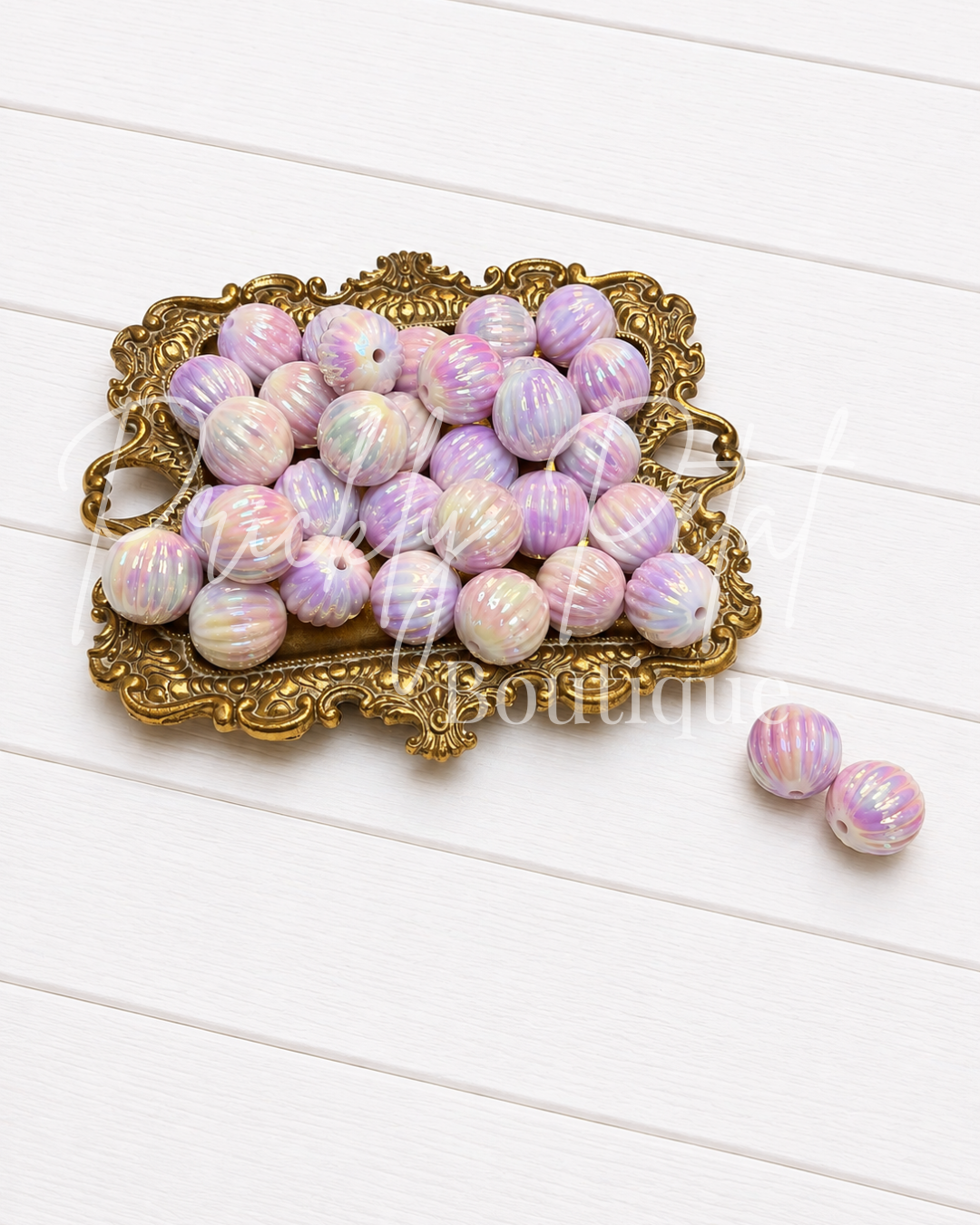 A group of round, iridescent beads with pink, purple, and white swirls, displayed on an ornate gold tray with two additional beads placed beside it on a white wood plank background.