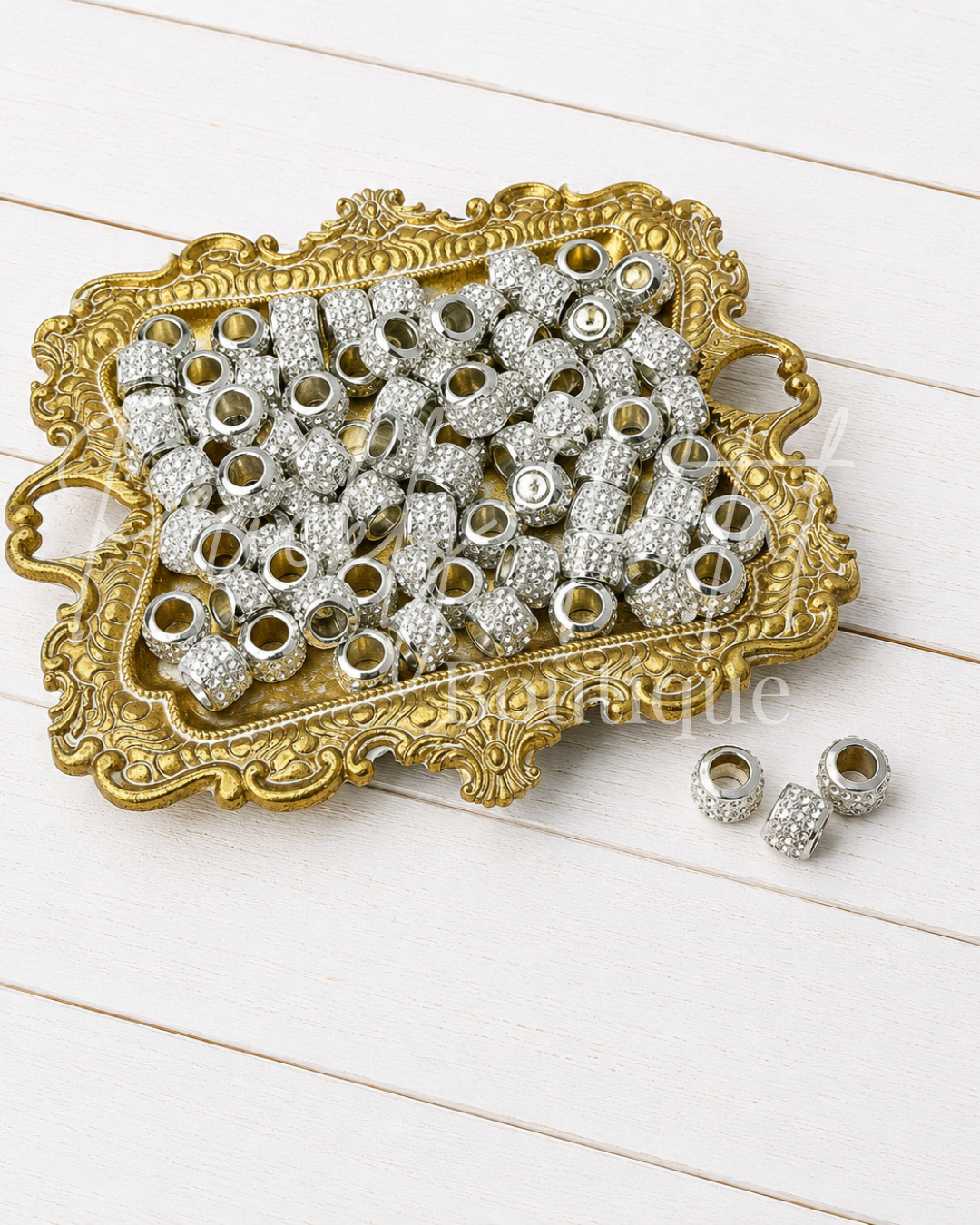 Silver rhinestone-embellished cylindrical and cylindrical-with-hole charms arranged on an ornate gold tray, with two additional charms placed beside the tray on a white wood background.