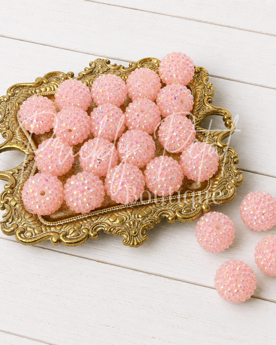 Pink rhinestone-covered round beads arranged on an ornate gold tray, with a few beads placed beside it on a white wood surface.