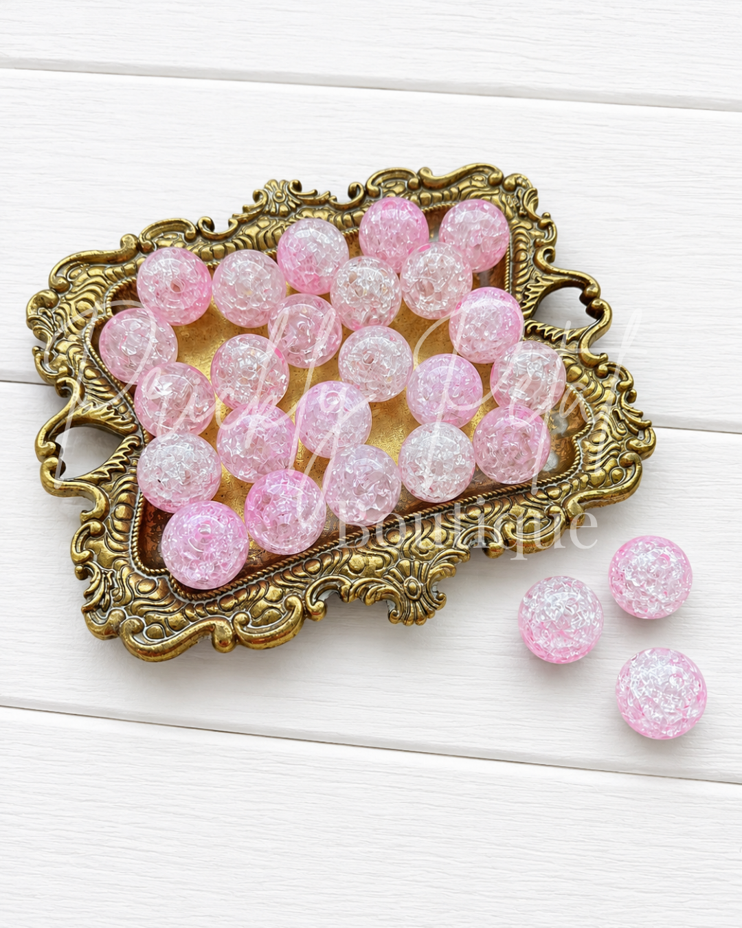 Pink and clear crackle glass beads arranged on an ornate gold tray, with three additional beads placed to the right, set against a white wood plank background.