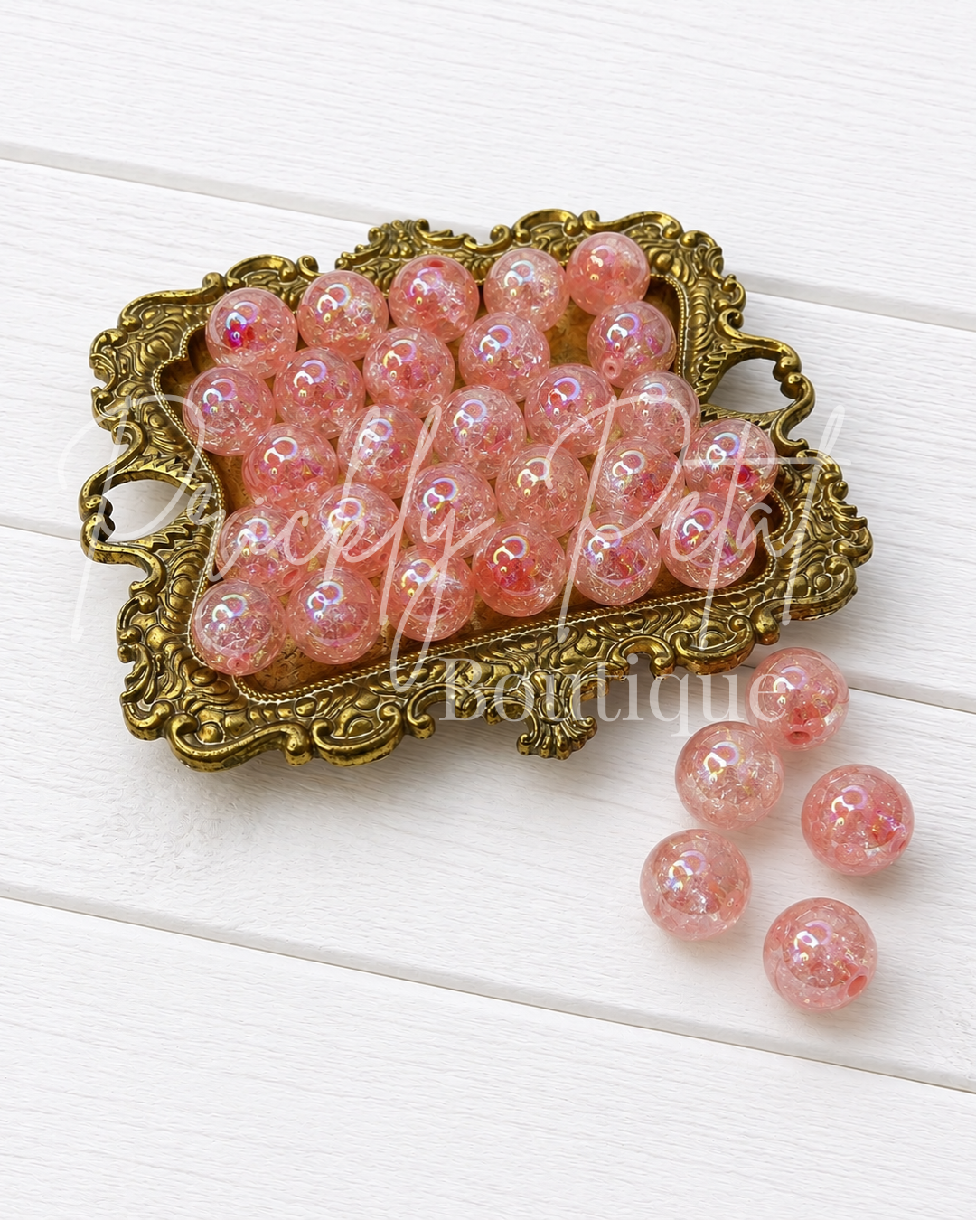 Pink glittery round beads displayed in an ornate gold-colored metal dish on a white wooden background, with four loose beads placed to the right.