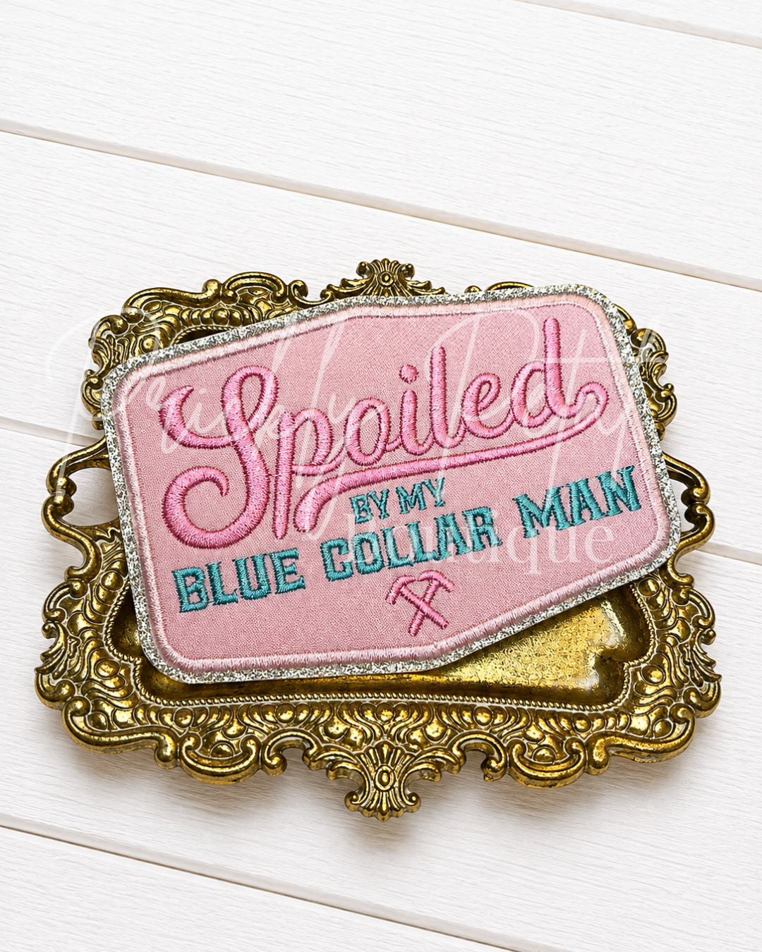 A decorative embroidered patch with a pink background and ornate gold border, reading 'Spoiled by my BLUE COLLAR MAN' in pink and teal lettering, placed on a white wood plank surface.