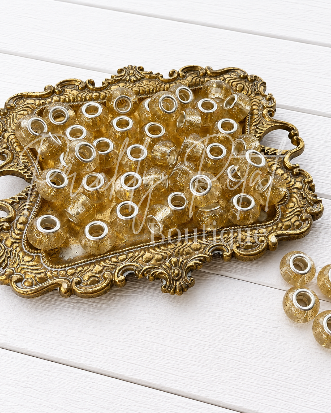 Clear resin craft beads with metallic gold swirls and silver metal eyelets, displayed on an ornate gold tray on a white wood background.
