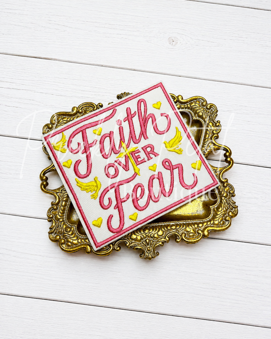 A square decorative sign with the phrase 'Faith Over Fear' in pink script on a white background, accented with small yellow hearts and birds, displayed on an ornate gold tray over a white wood plank surface.