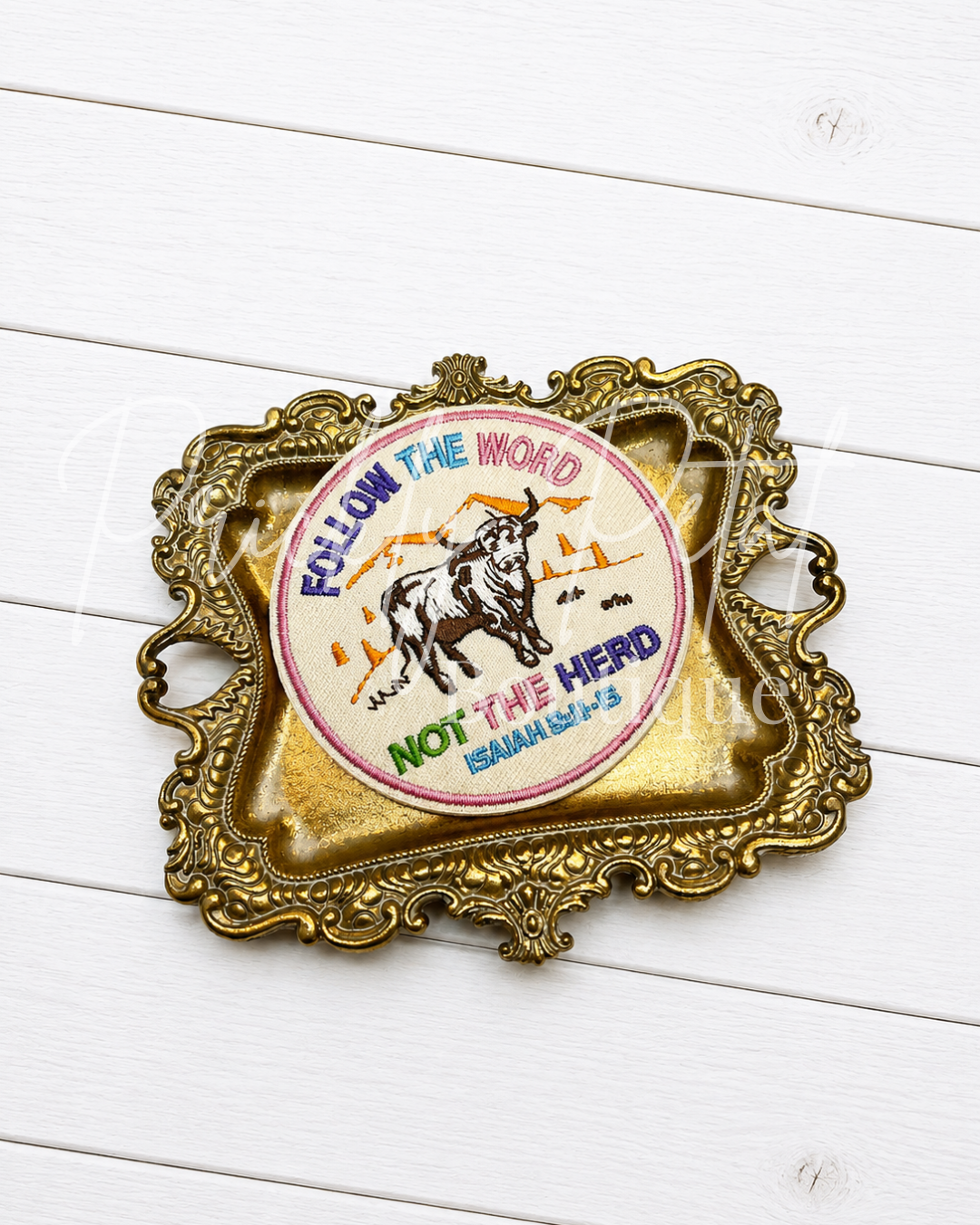 A decorative gold-toned ornate tray featuring a circular vintage-style patch illustration with a bull and the text 'FOLLOW THE WORD NOT THE Herd' on a white wood plank background.