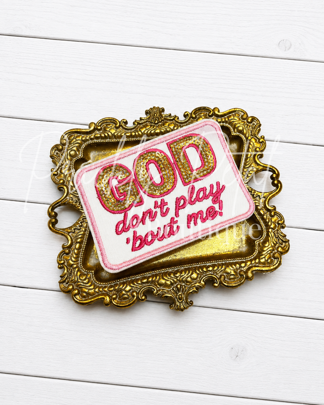 A decorative embroidered patch with the text 'GOD don't play bout me!' in pink and gold, presented on an ornate gold frame over a white wood plank background.