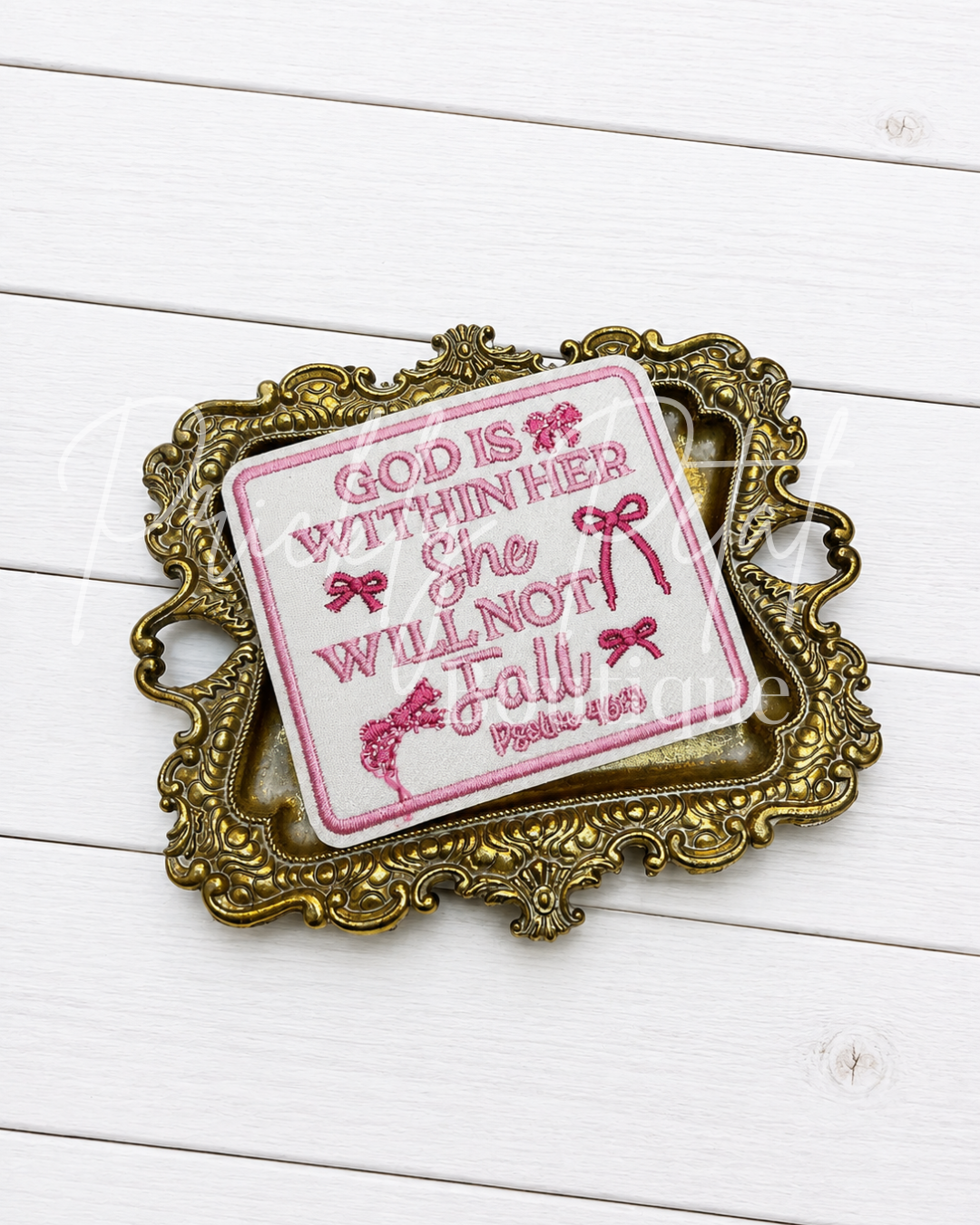 An embroidered patch with the Bible verse 'God is within her She will not Fall' in pink on a white background, styled inside an ornate gold-colored frame on a white wood plank surface.