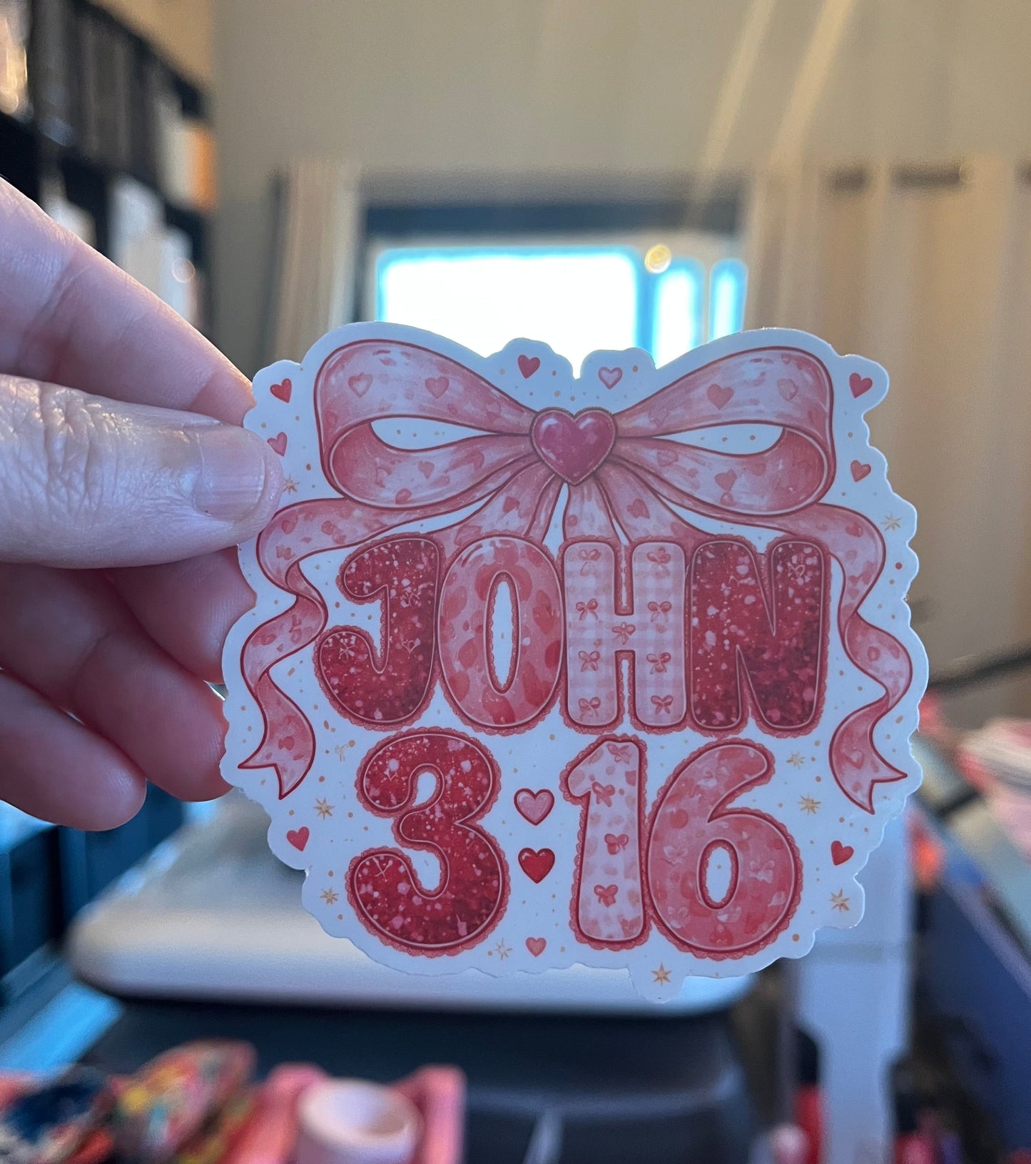 Sticker with 'JOHN 3:16' text and decorative elements held by a hand.