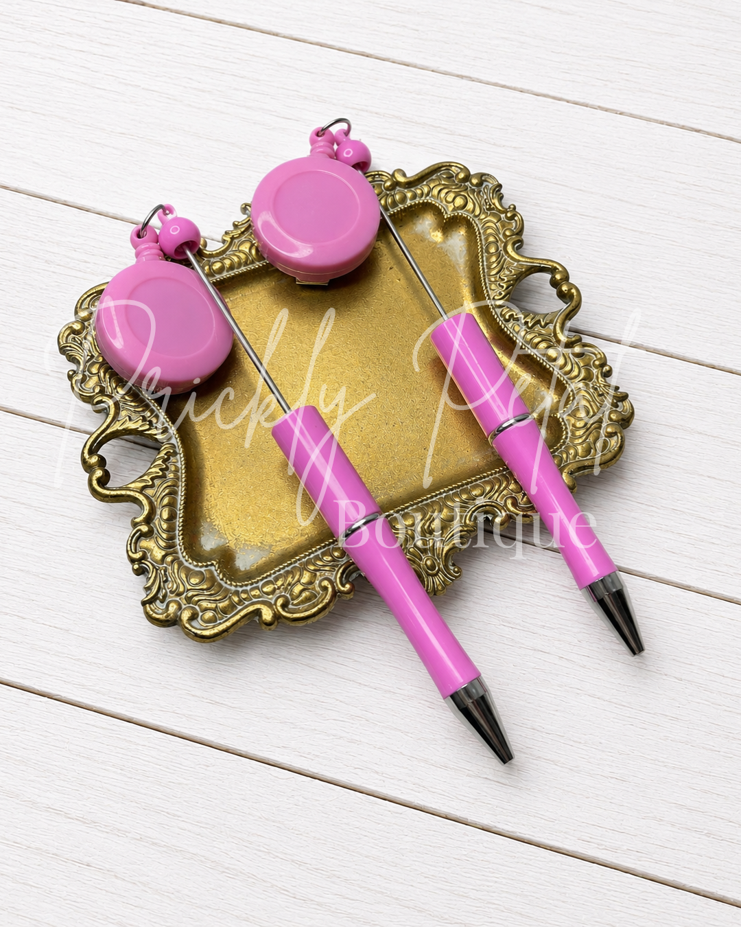 Two pink pen-style tools with round pink ends and metal tips, arranged on an ornate gold tray over a white wood background.