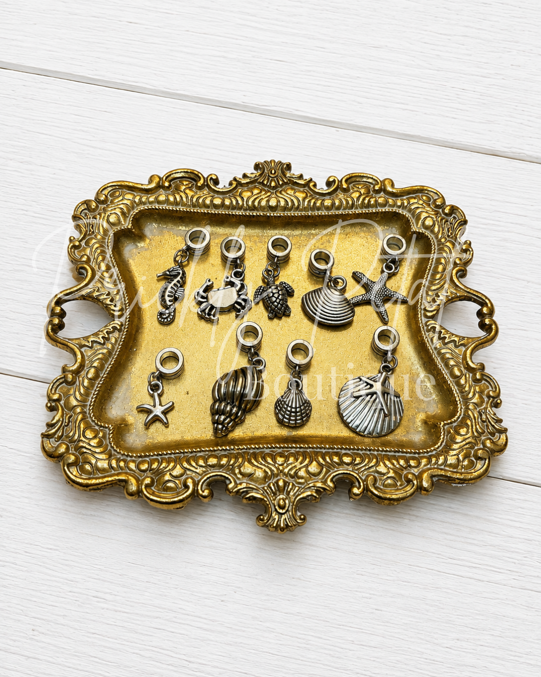 A set of nine silver-toned nautical-themed jewelry charms, including seashells, starfish, and a seahorse, displayed on an ornate gold-colored metal tray over a white wood background.