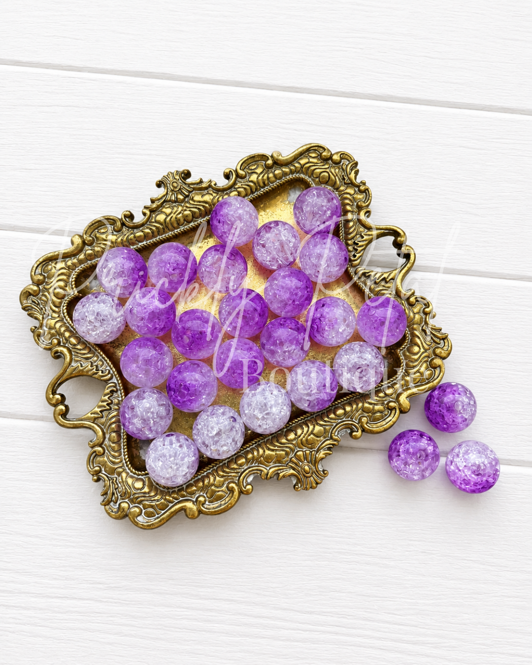 Purple and lavender round beads with a glittery, crackled texture, displayed in an ornate gold-colored metal tray on a white wooden background.