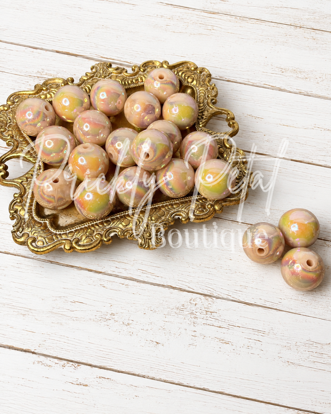 Round marbled beads in pastel pink, peach, and yellow tones, displayed on an ornate gold tray with three additional beads on a white wood plank background.