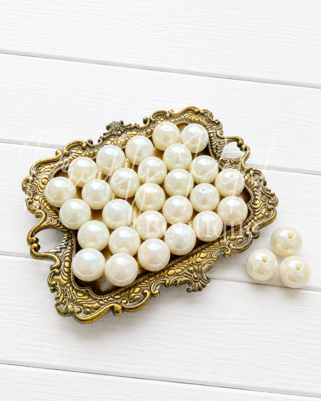 A group of round, glossy white faux pearls arranged on an ornate antique gold tray, with three additional pearls placed beside it on a white wooden surface.