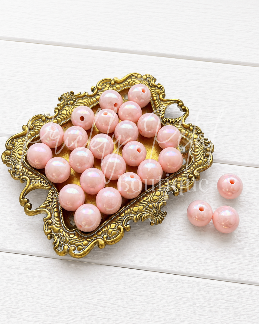 Pink round craft beads with holes, displayed in an ornate gold-colored metal dish on a white wooden background, with three additional beads placed to the side.