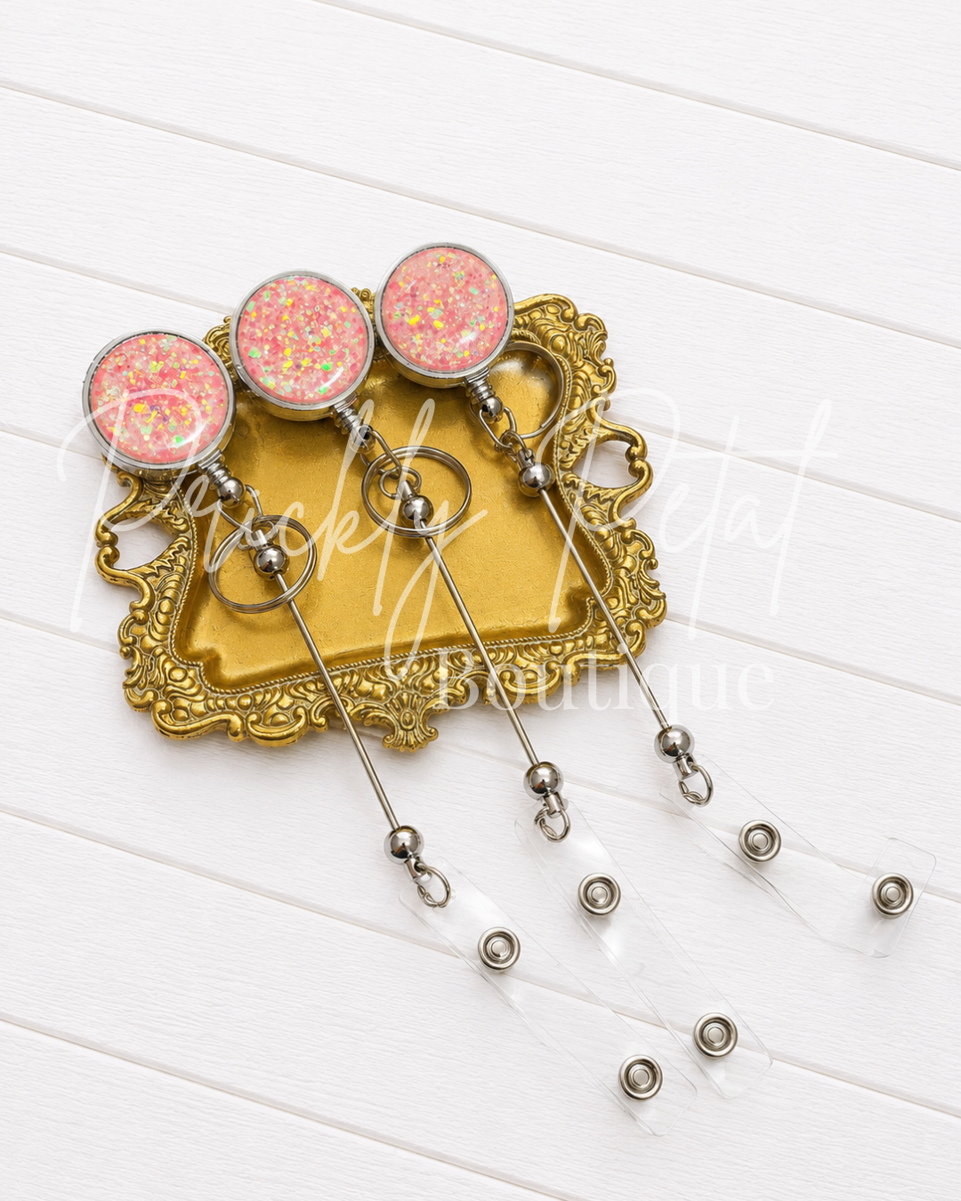 Three pink glitter-covered ID badge holder stems with round silver-rimmed tops, displayed on an ornate gold tray over a white wood plank background.