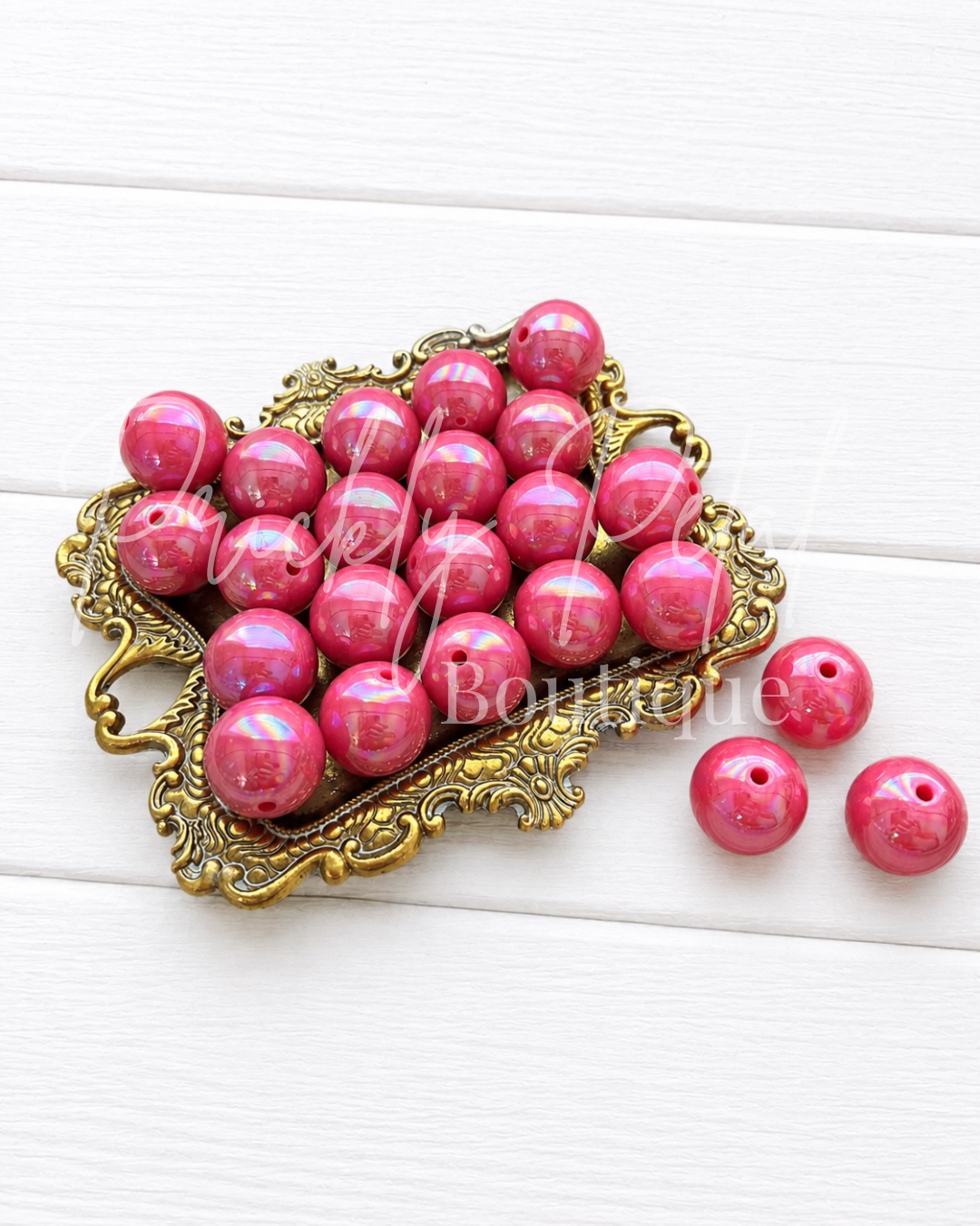 A pile of glossy pink round beads with a small hole, displayed on an ornate gold-colored metal tray with three beads placed beside it on a white wood background.