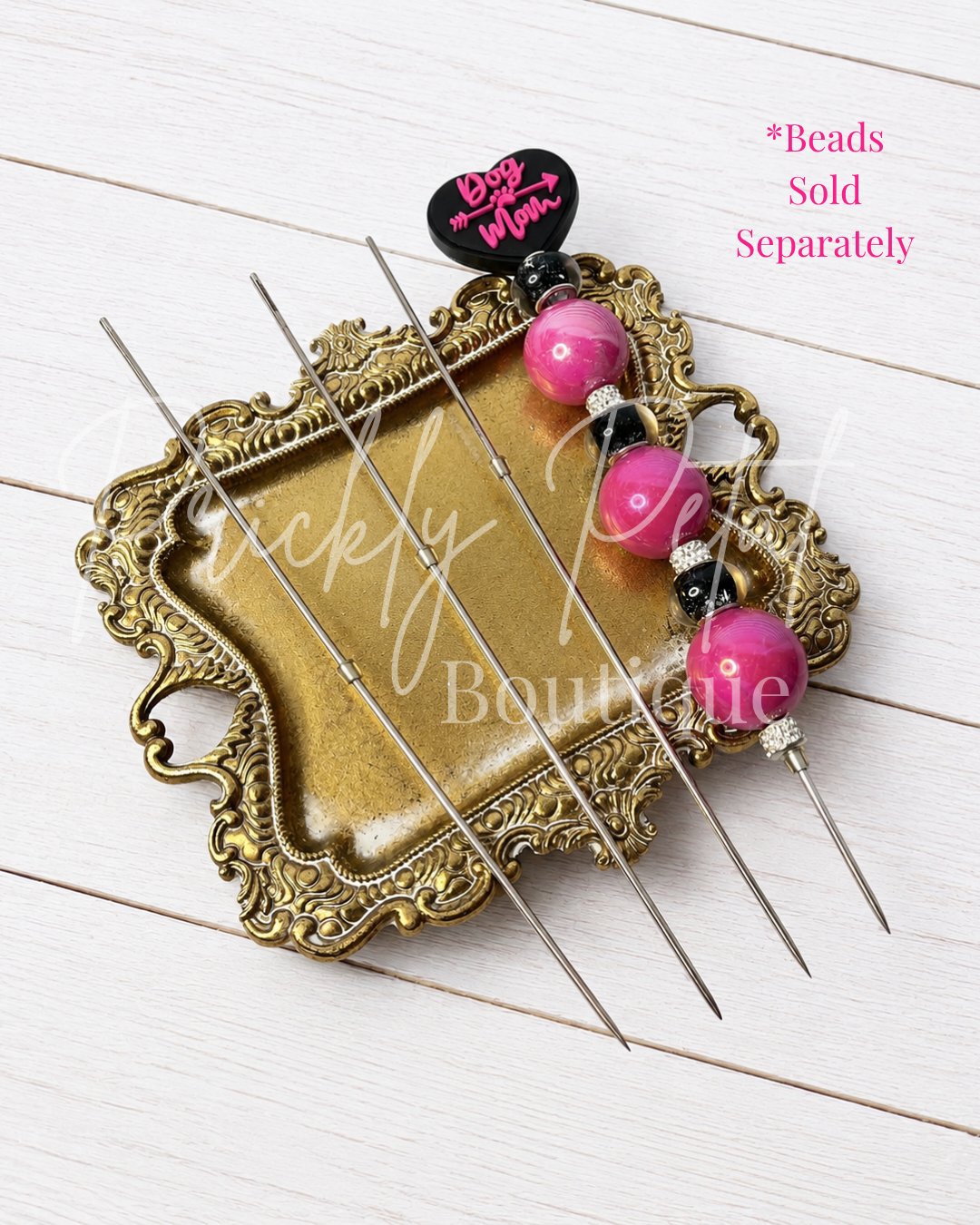 A set of metal bead wire picks with pink and black beads, displayed on an ornate gold tray over a white wood background.