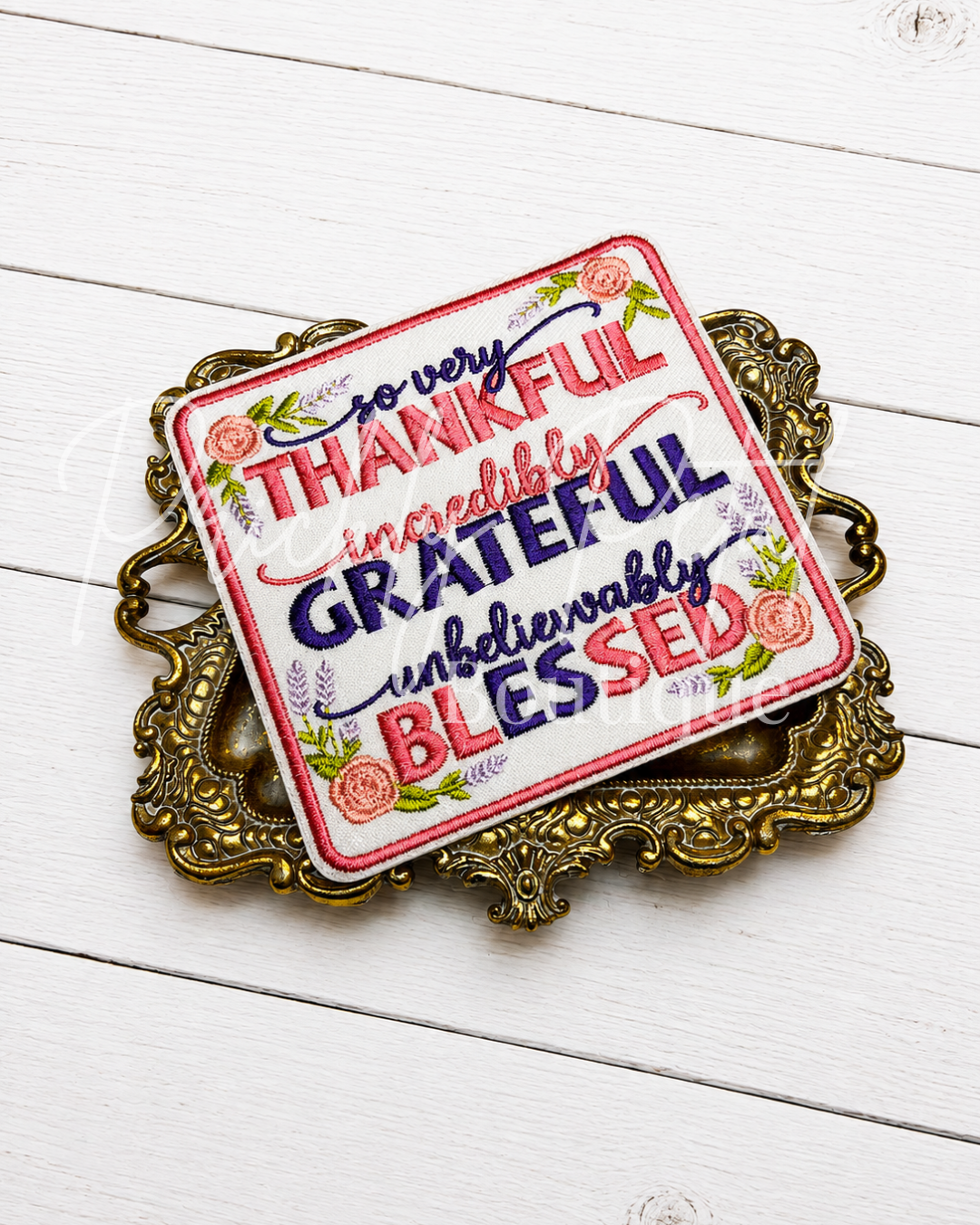 A square decorative sign with floral and script text reading 'so very THANKFUL incredibly GRATEFUL unbearably BLESSED,' displayed on an ornate gold tray over a white wood plank surface.