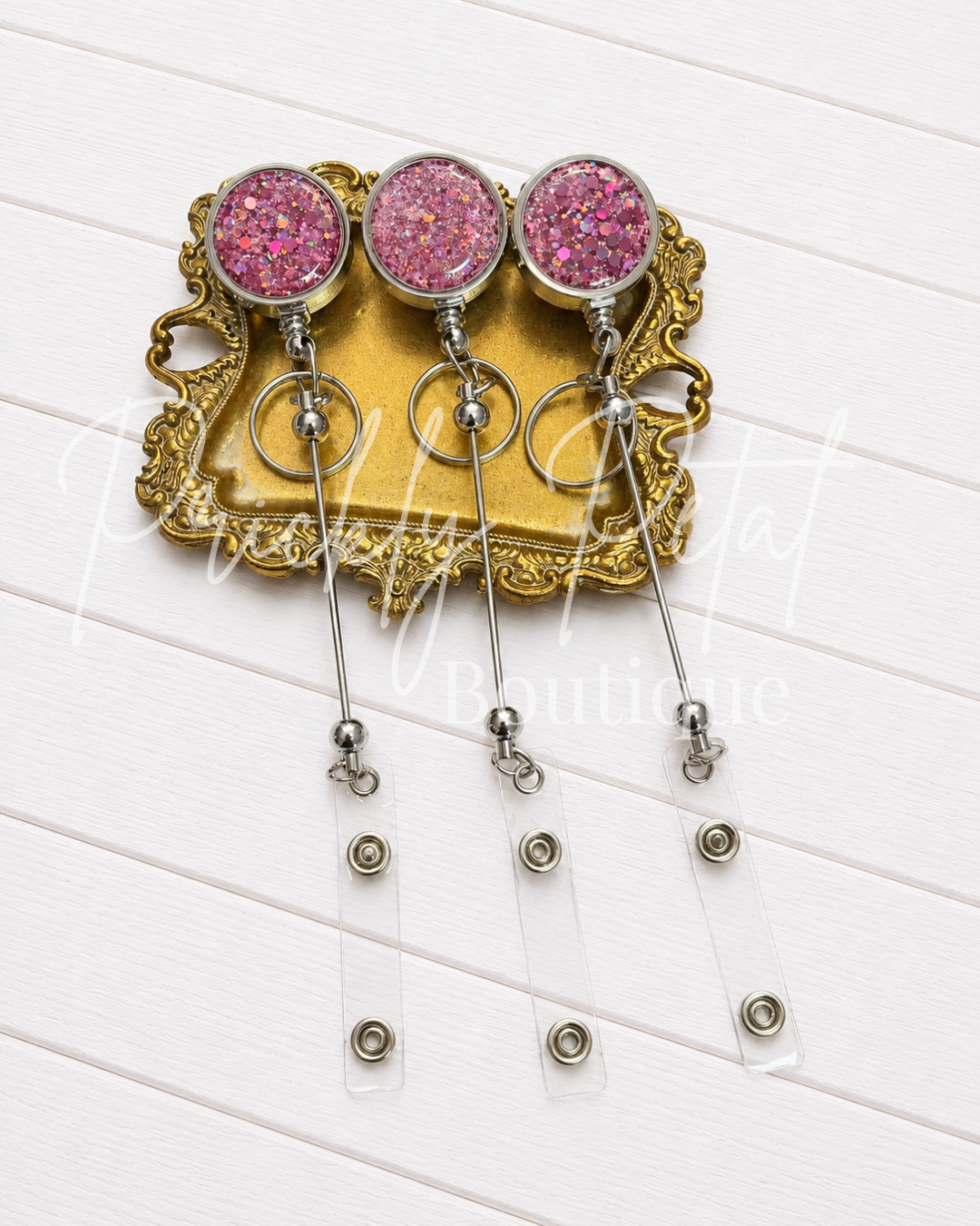 Three pink glitter ball-beaded retractable badge reels with silver metal rods and clear plastic pull tabs, displayed on an ornate gold tray over a white wood plank background.