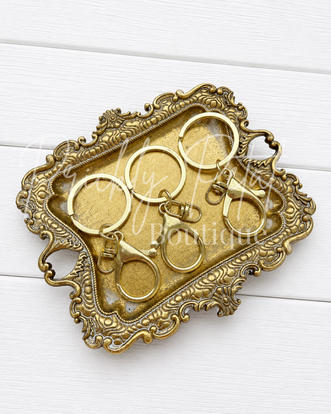 An ornate antique-style gold tray with a decorative carved border, holding several gold key rings and keychains on a white wood plank background.