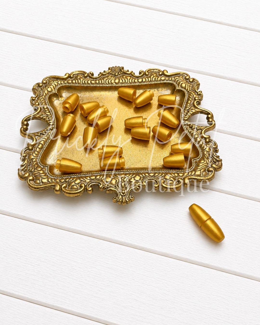 Gold metallic bullet casings displayed in an ornate gold tray on a white wood plank background, with one casing placed outside the tray.