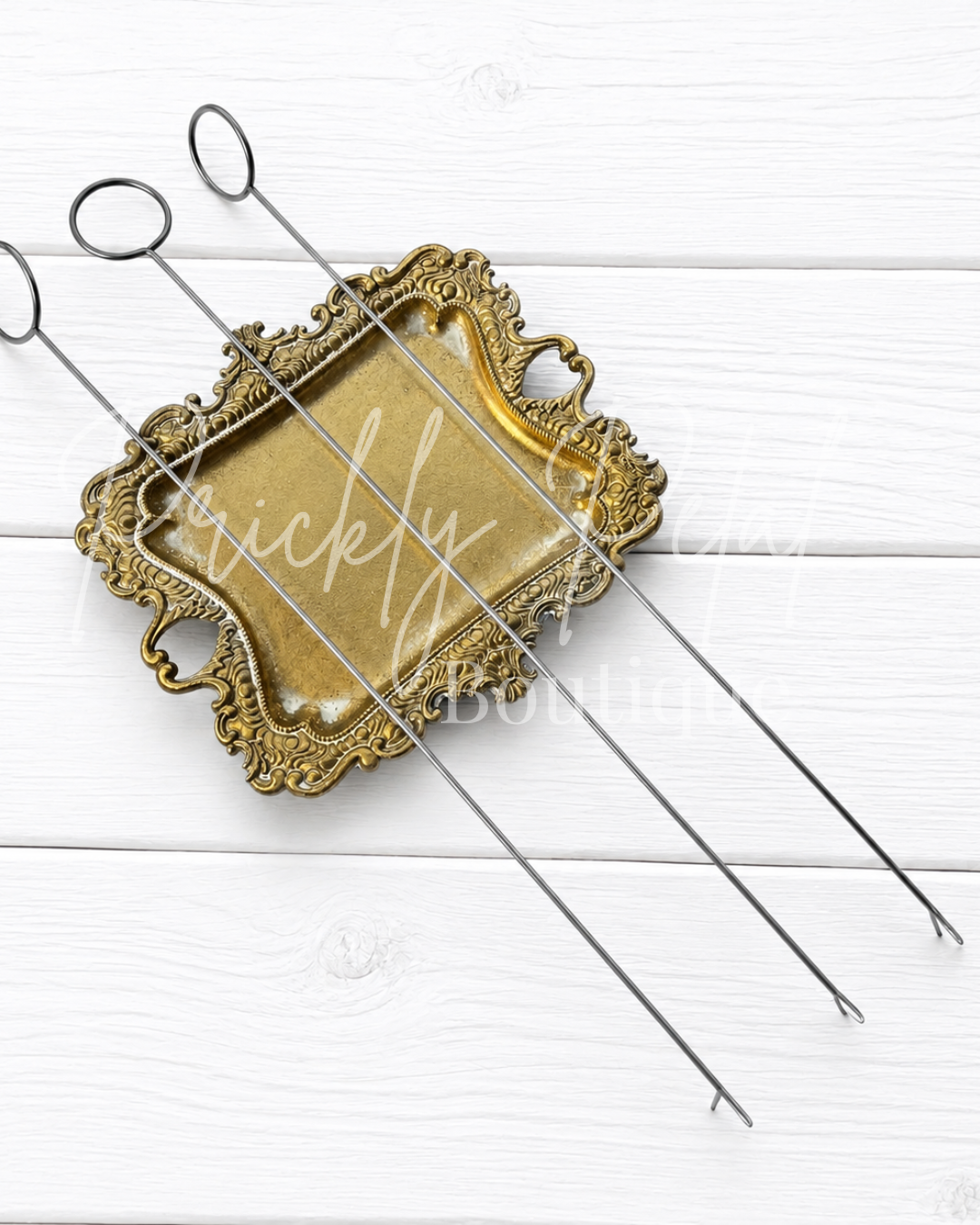 Three metal skewer tools with looped handles rest on an ornate gold-colored decorative tray over a white wood plank background.