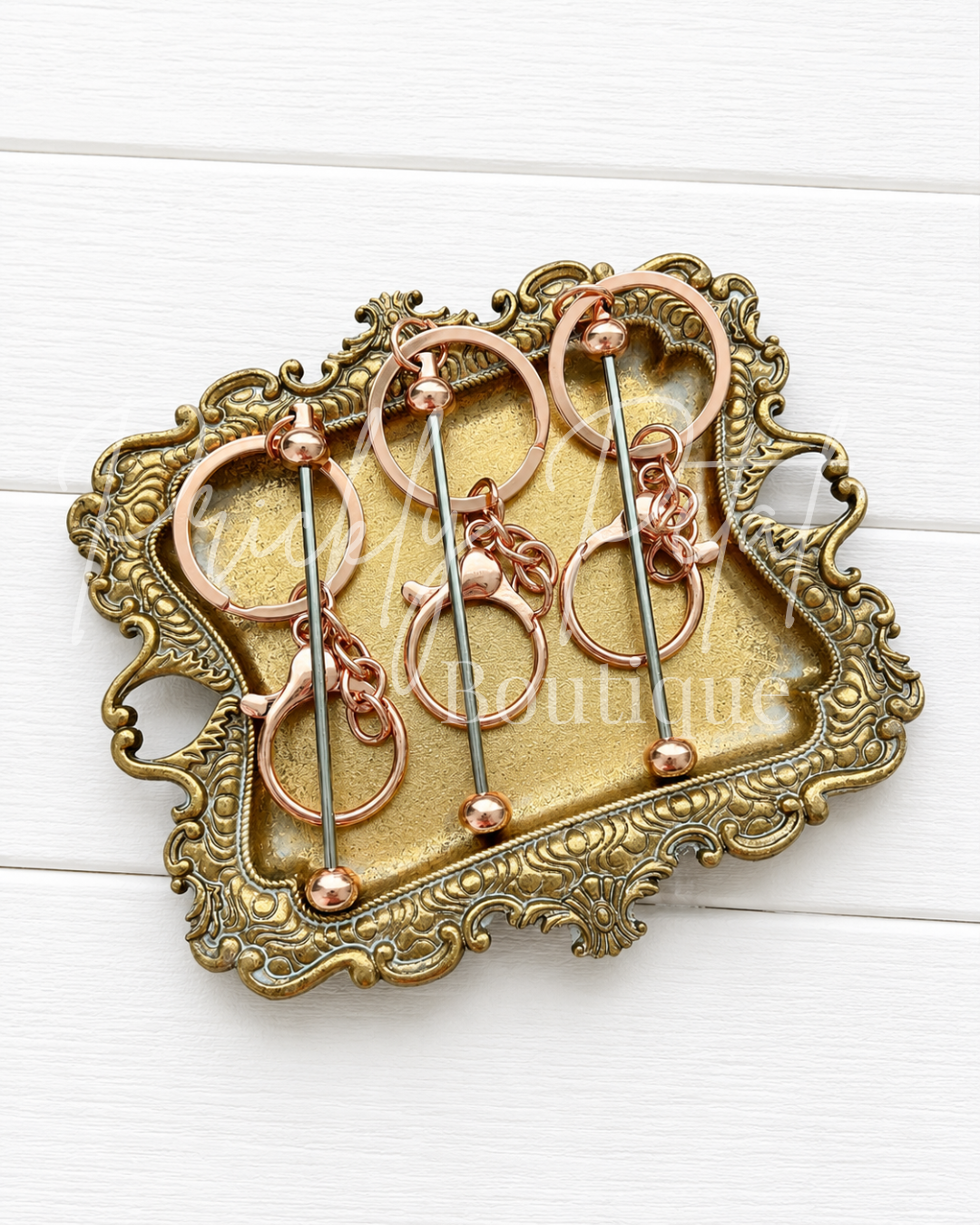Three rose gold and silver beadable bar keychains displayed on an ornate vintage-style gold tray over a white wood background.