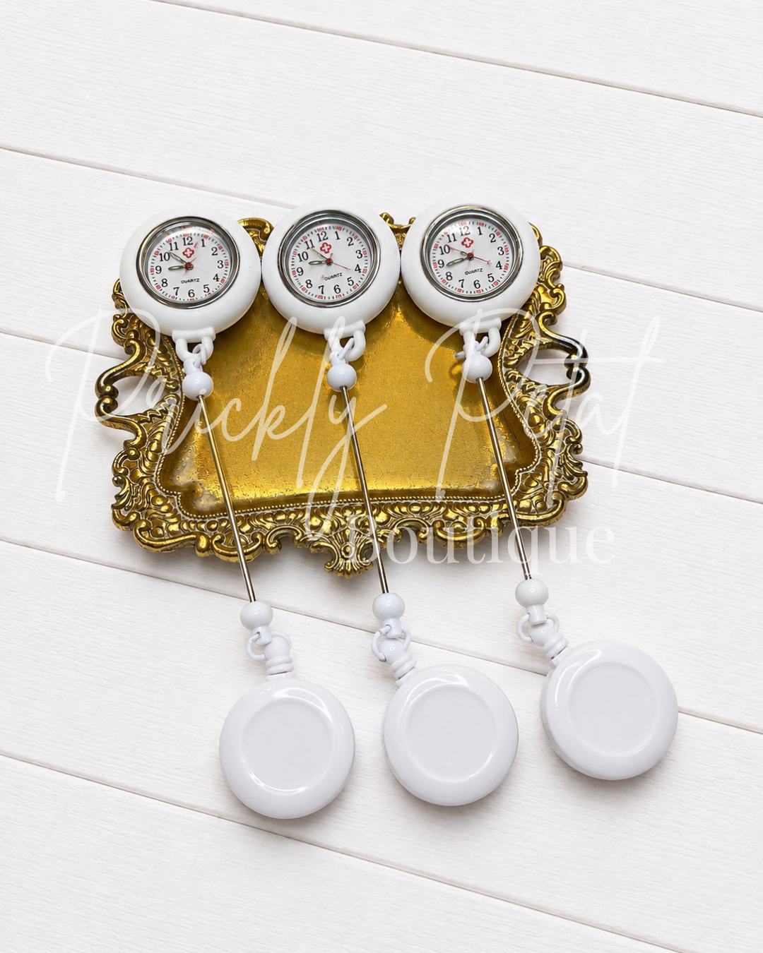 Three white analog pocket watches with round dials and long metal chains, arranged on an ornate gold tray over a white wood plank background.