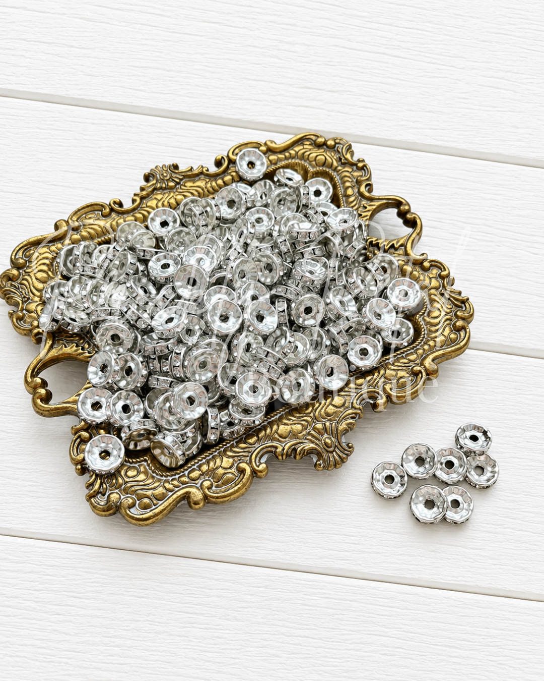 Silver metal spacers with central holes, shown piled in an ornate gold tray with a few placed beside it on a white wood background.