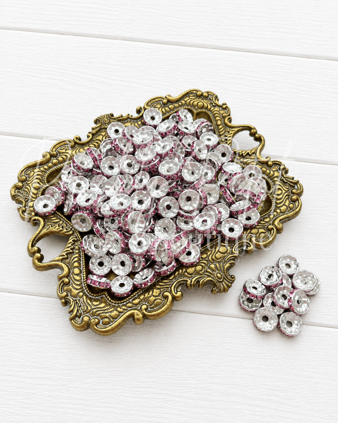 A decorative gold-toned tray filled with small round clear rhinestone beads with pink glitter trim, alongside a few loose beads on a white wood background.