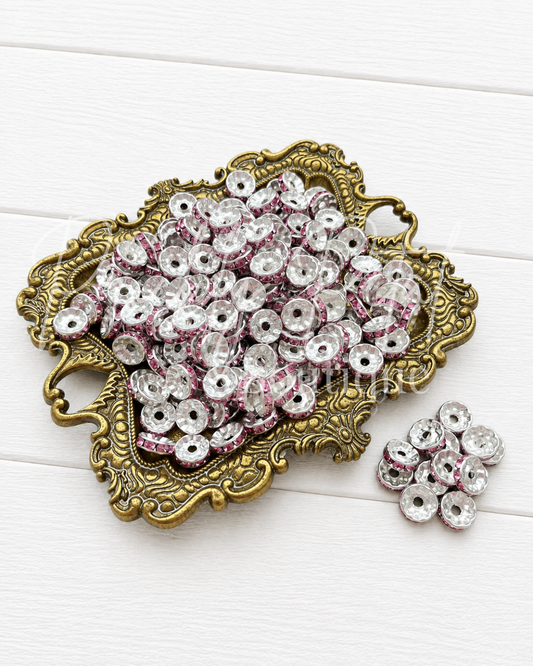 A decorative gold-toned tray filled with small round clear rhinestone beads with pink glitter trim, alongside a few loose beads on a white wood background.