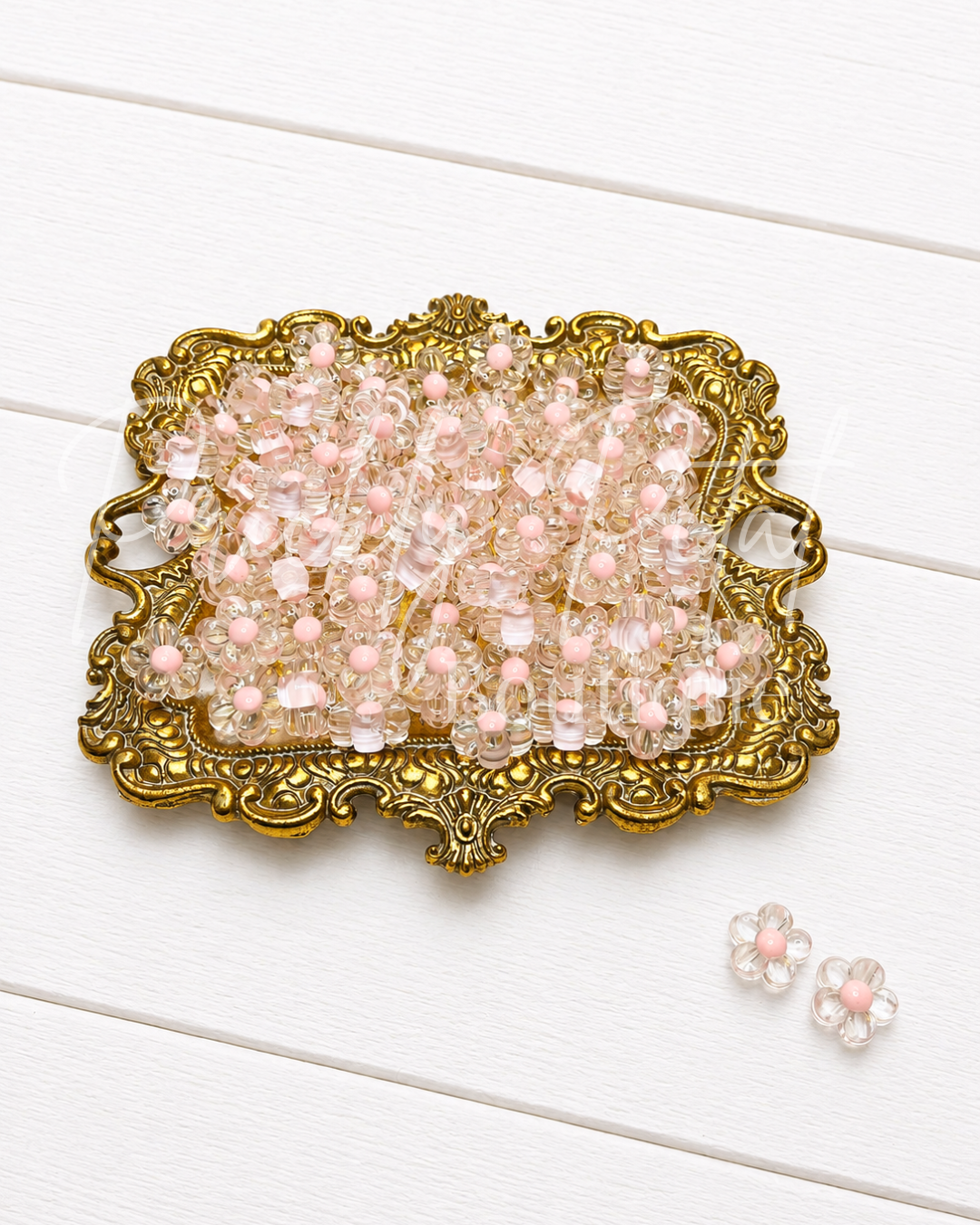 Clear crystal beads with pale pink centers arranged in an ornate gold tray on a white wood background, with two matching flower-shaped beads placed nearby.