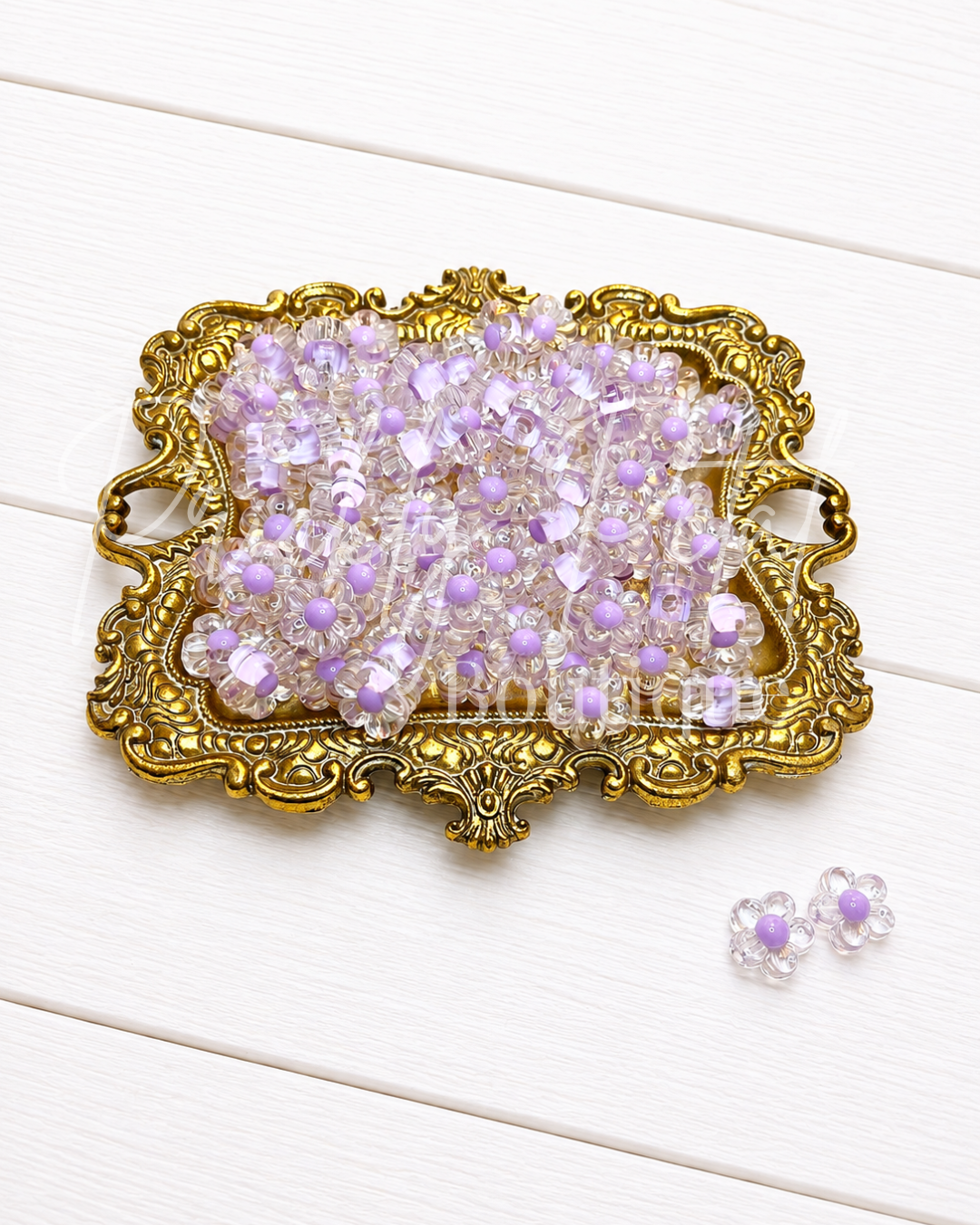 A decorative gold tray filled with small clear rhinestone flower-shaped beads with light purple centers, with two matching loose beads placed below on a white wood plank background.