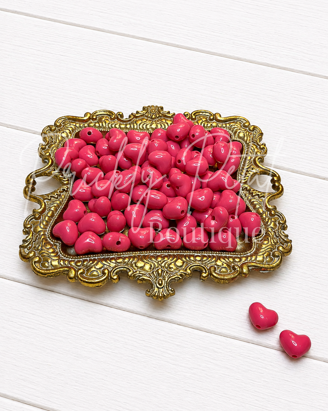 Bright pink heart-shaped beads arranged in an ornate gold tray on a white wood background, with two loose beads placed below the tray. 