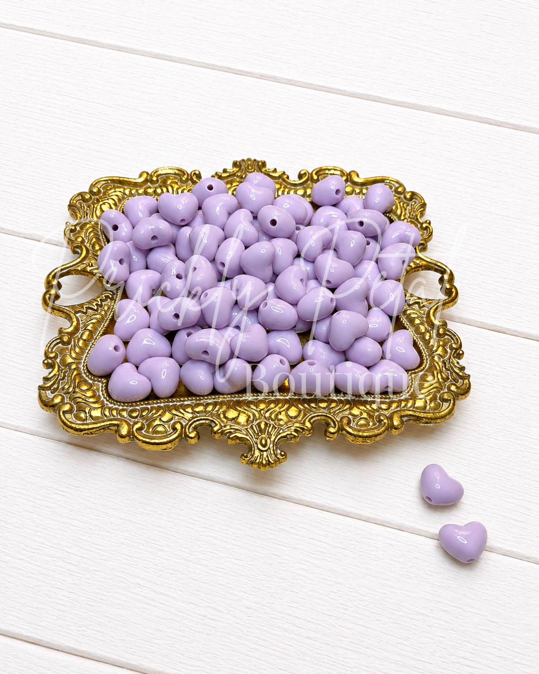 Lavender heart-shaped wooden beads with holes, displayed in an ornate gold-colored tray on a white wooden background.