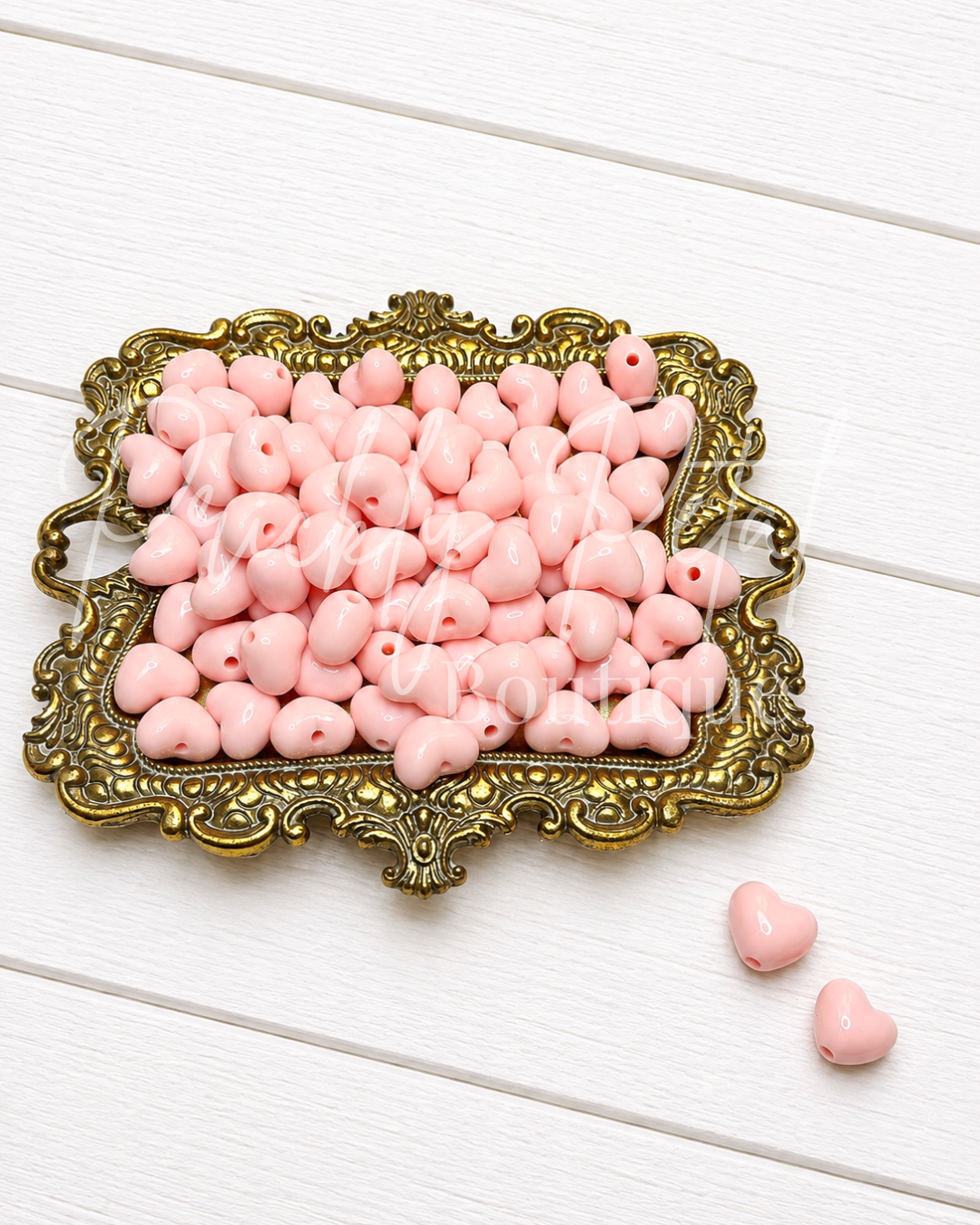Pink heart-shaped wooden beads with holes, displayed on an ornate gold tray with two additional beads on a white wood background.