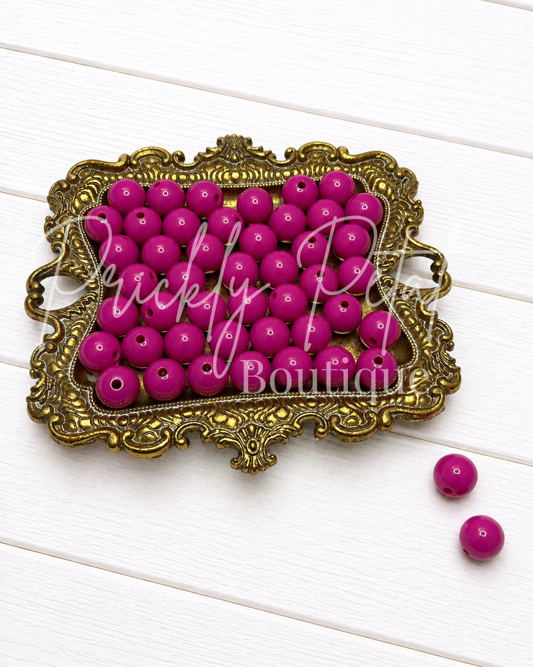 Bright pink wooden bead blanks arranged in an ornate gold-colored metal tray on a white wood plank background, with two loose beads placed to the right. 