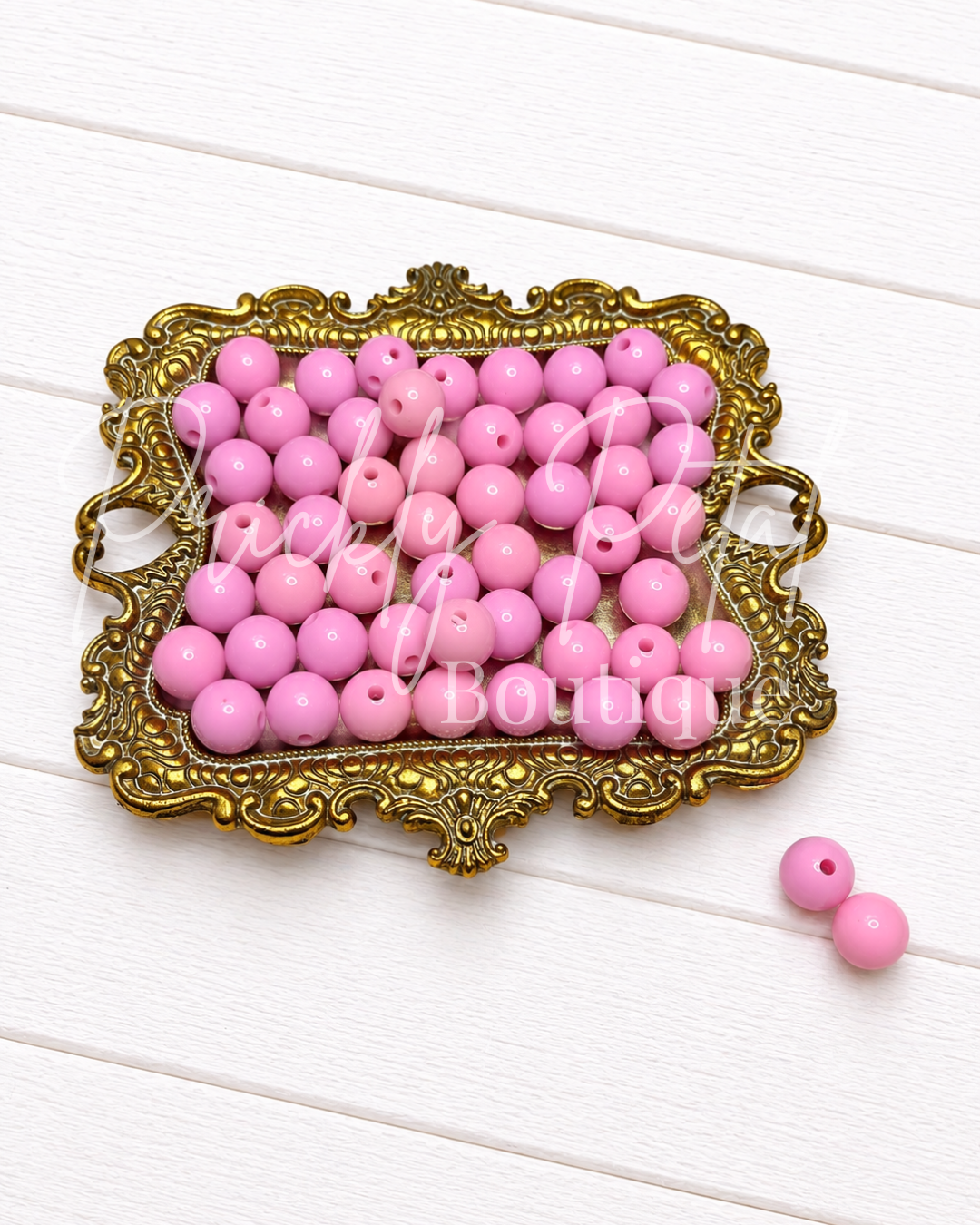 Pink round craft beads with holes, displayed in an ornate gold-colored frame on a white wooden background, with two loose beads in the bottom right corner.