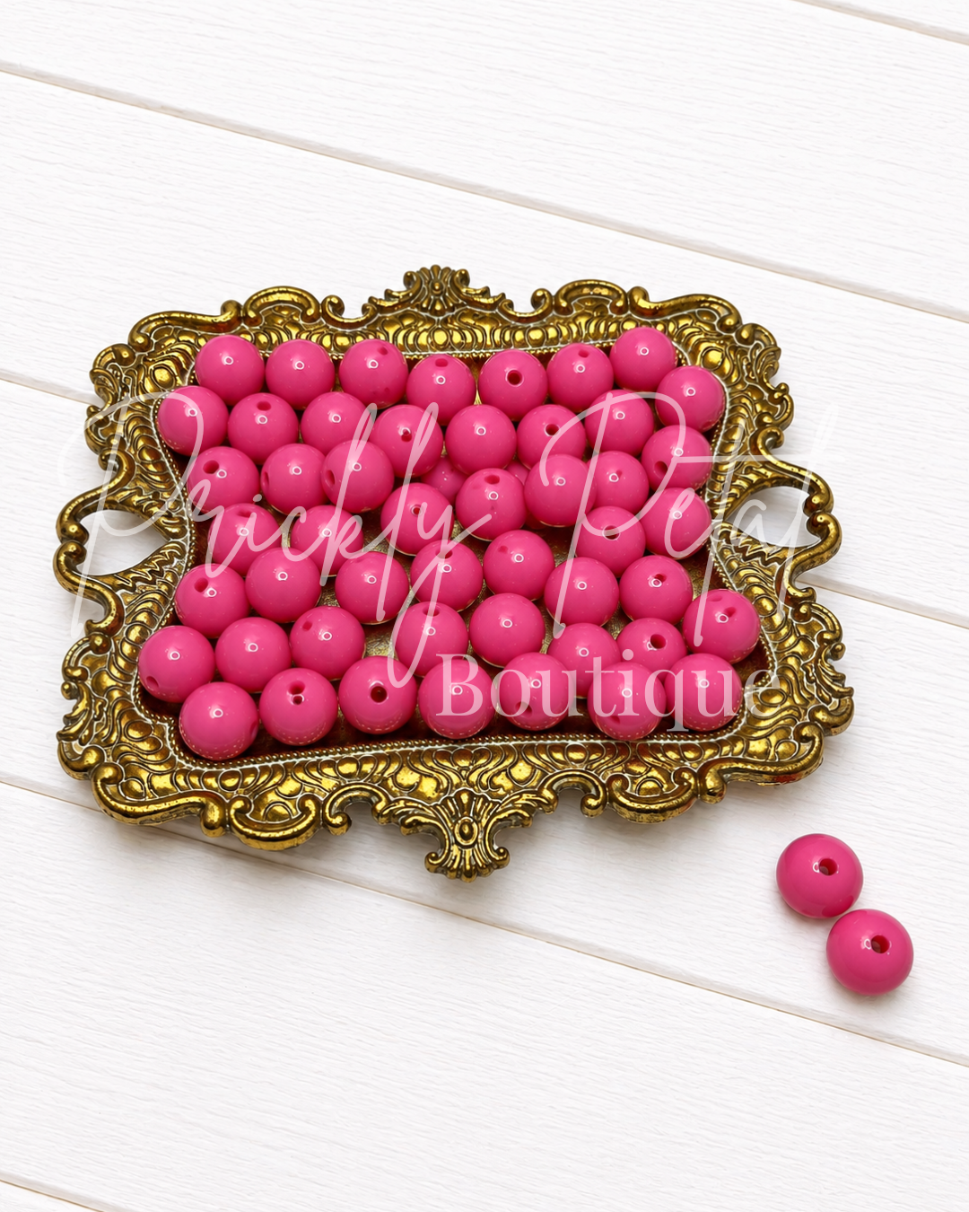 Bright pink wooden beads with drilled holes, displayed on an ornate gold tray with two additional beads on the side, set against a white wood plank background.