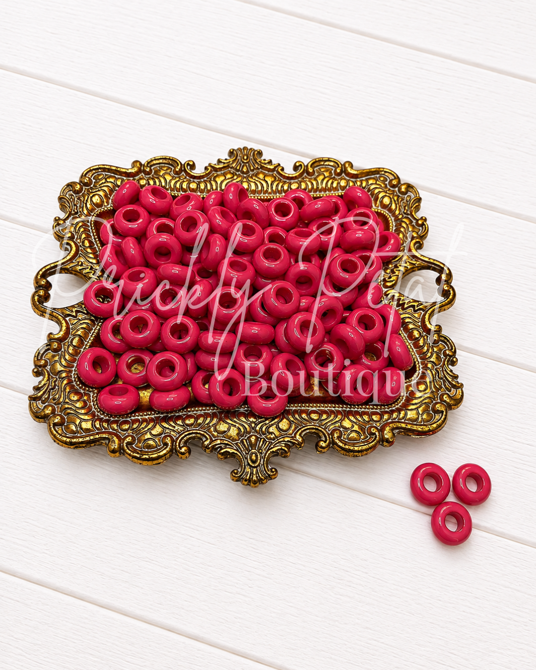 Bright pink plastic seed beads arranged in an ornate gold-colored decorative tray, with three additional beads placed to the side on a white wood plank background. 