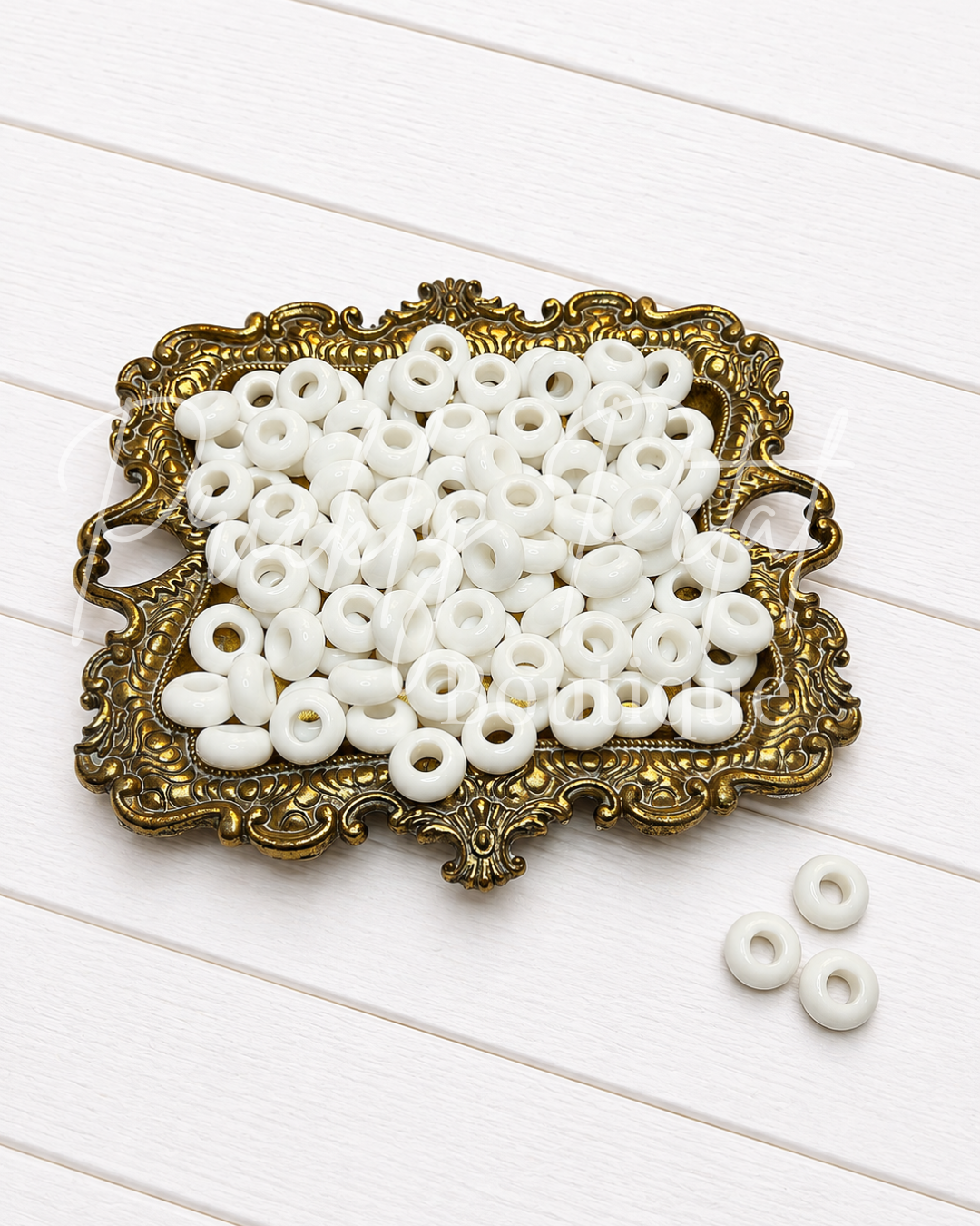 White round spacer beads with center holes, displayed in an ornate gold tray on a white wood background.