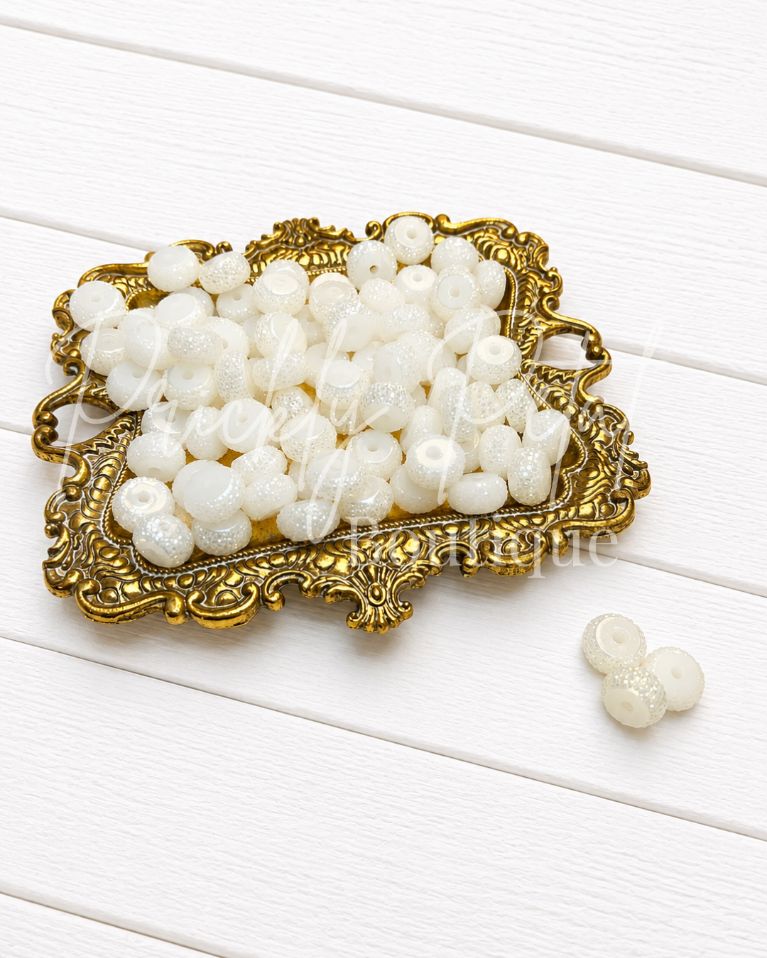 White round glass beads with a glittery finish, displayed on an ornate gold-colored tray with a few loose beads on a white wooden background.