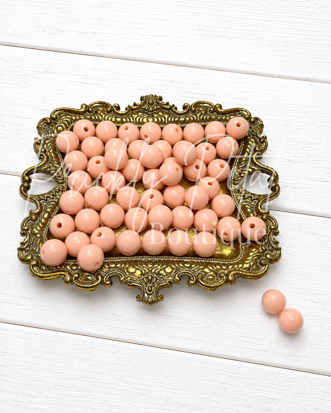 A tray of peach-colored round beads with two loose beads placed beside it on a white wooden surface. The beads are displayed in an ornate gold-colored metal dish, 