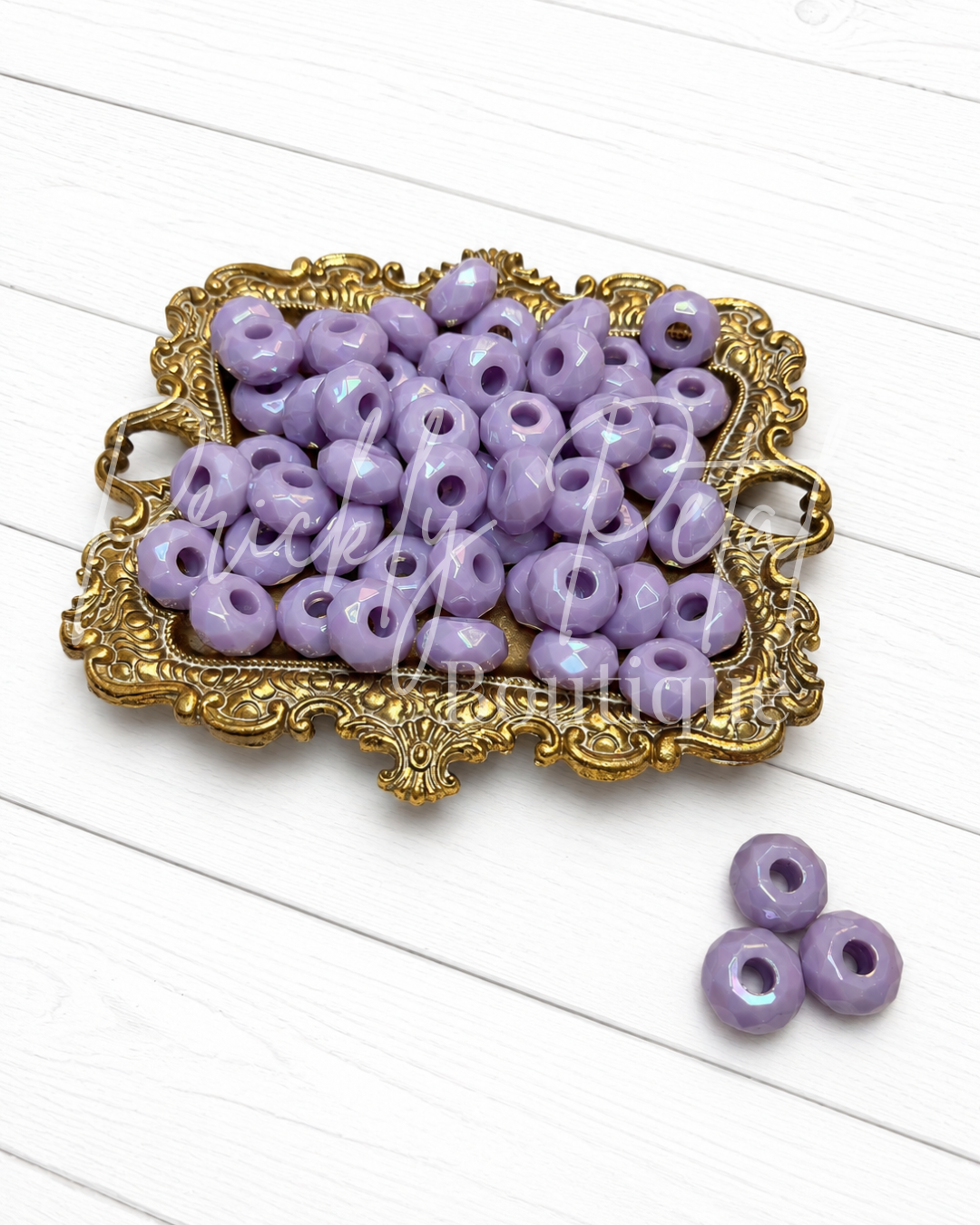 Lavender purple round craft beads with center holes, displayed in an ornate gold tray with three additional beads on a white wood background.