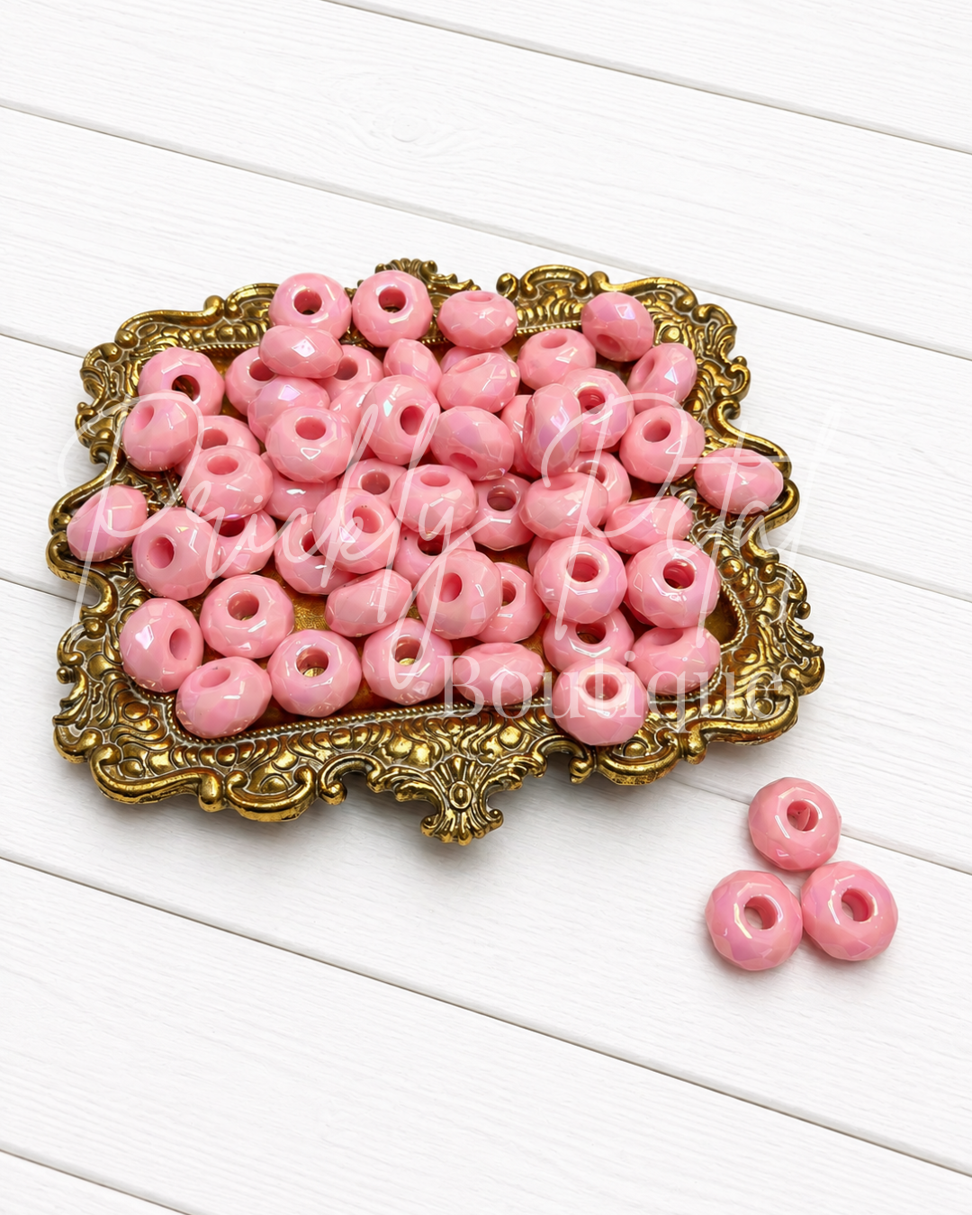 Pink round acrylic beads with center holes, displayed on an ornate gold tray with three additional beads on the side, set against a white wooden plank background.